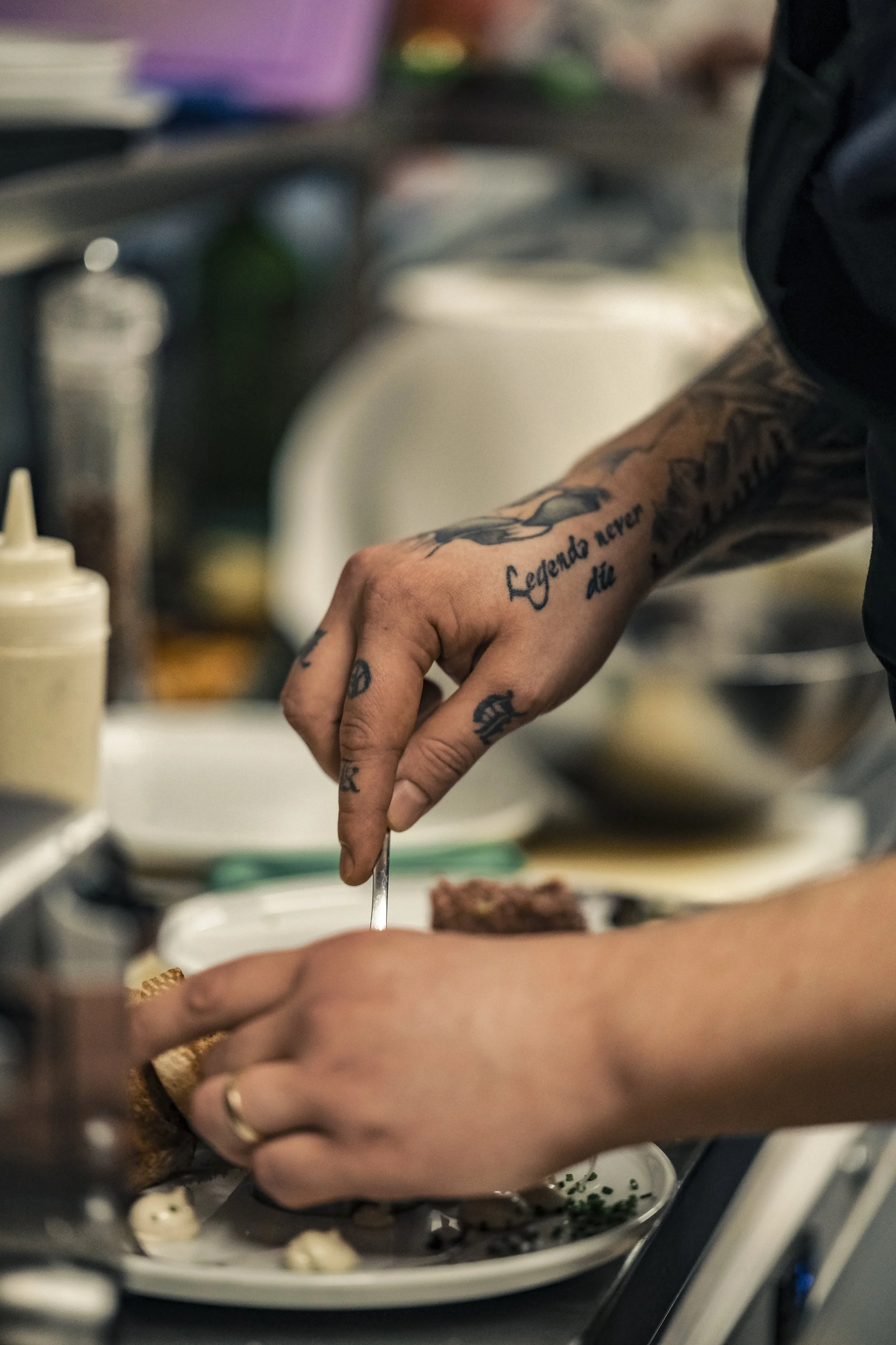 A person with tattooed arms preparing food, using a utensil to place ingredients on a white plate in a kitchen.