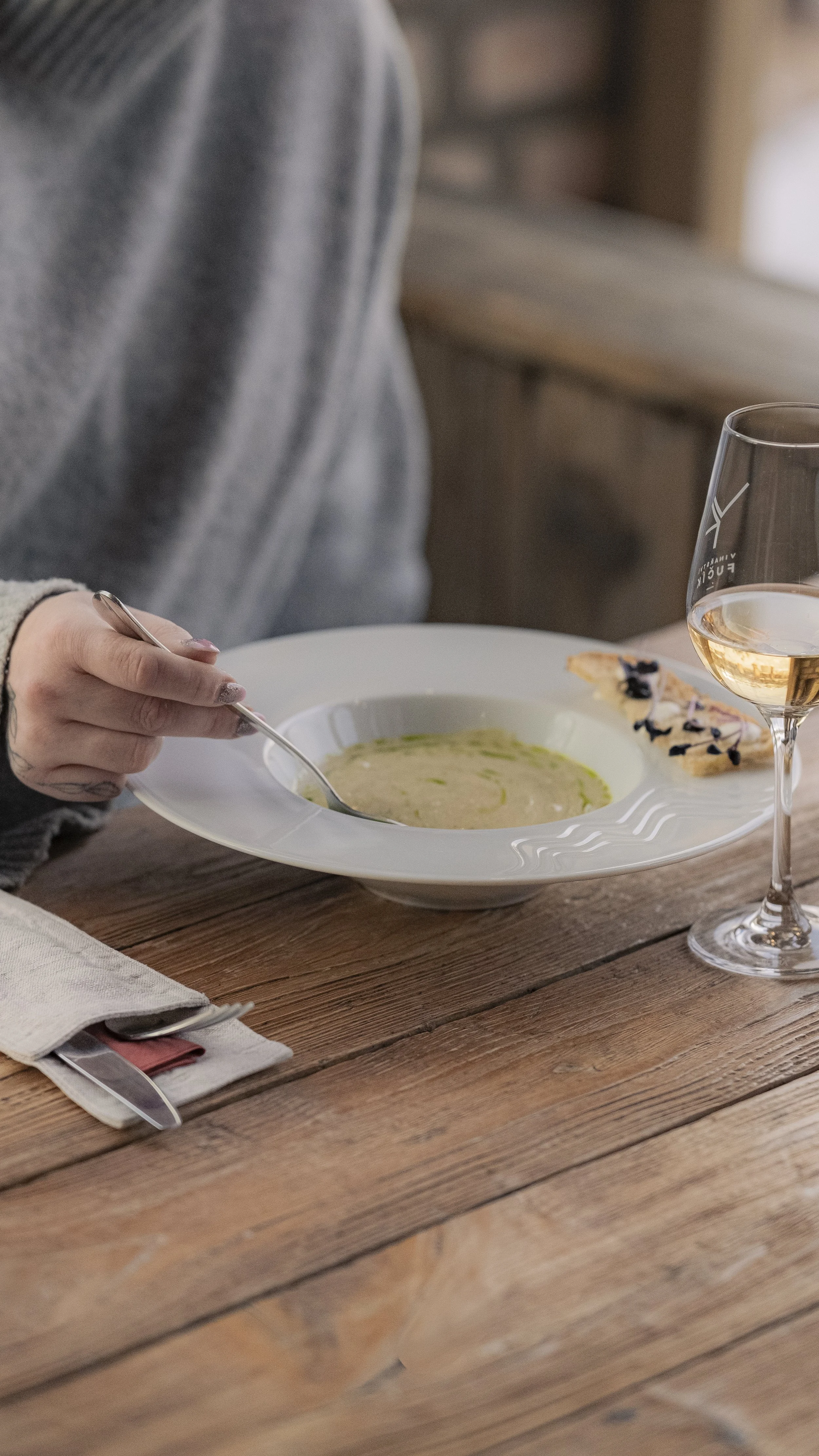 A person sitting at a wooden table, holding a spoon over a bowl of soup, with a glass of white wine and a piece of bread with berries nearby.