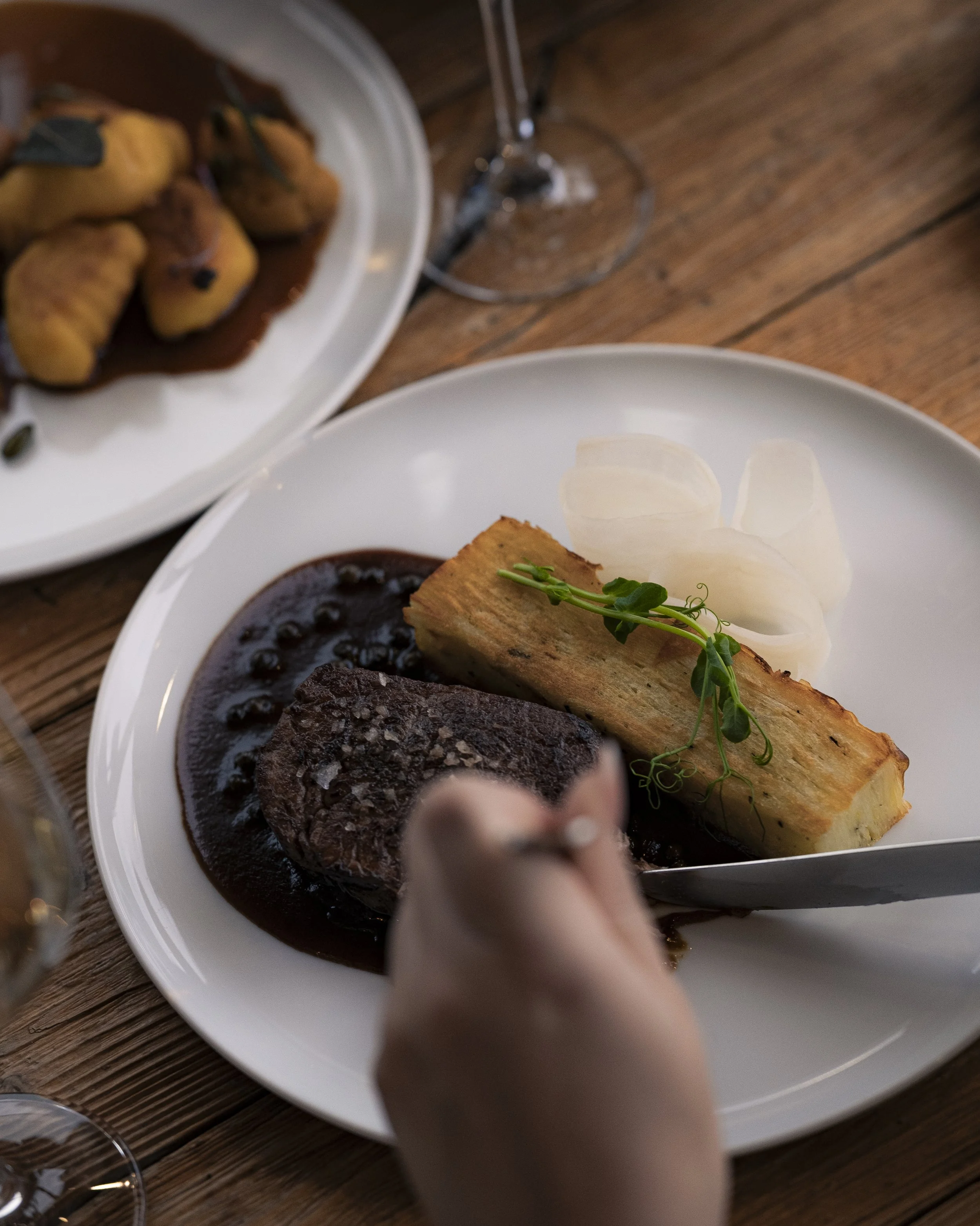 A plated meal with beef stew, mashed potatoes with onion slices, and a piece of fried fish garnished with microgreens, on a wooden table with a glass of wine nearby.