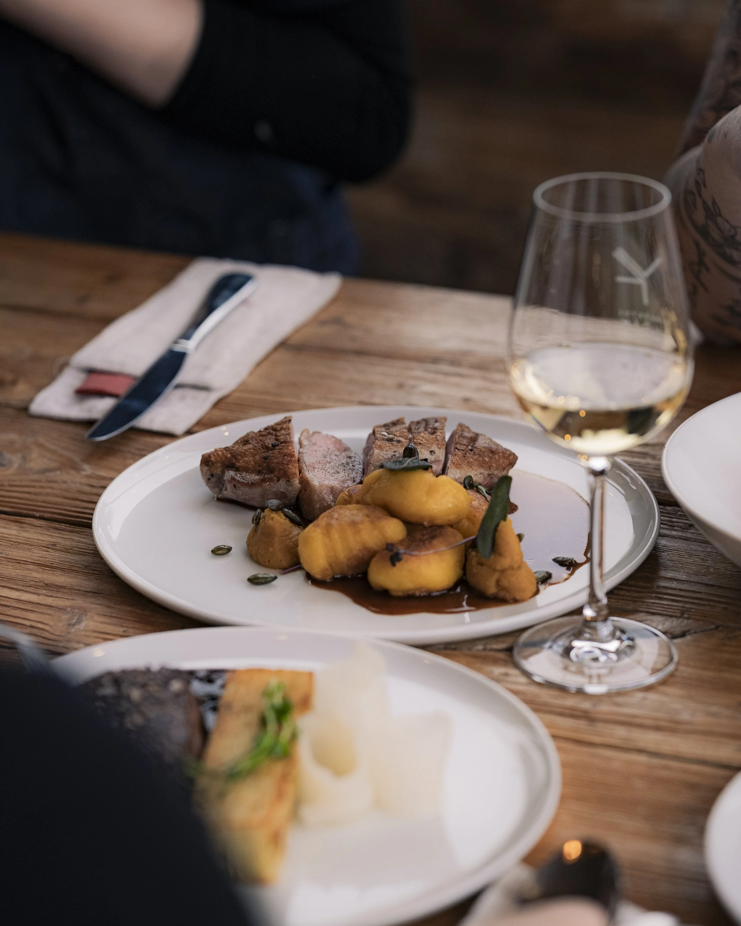 A plated meal with slices of meat and yellow vegetables, accompanied by a glass of white wine on a wooden table.