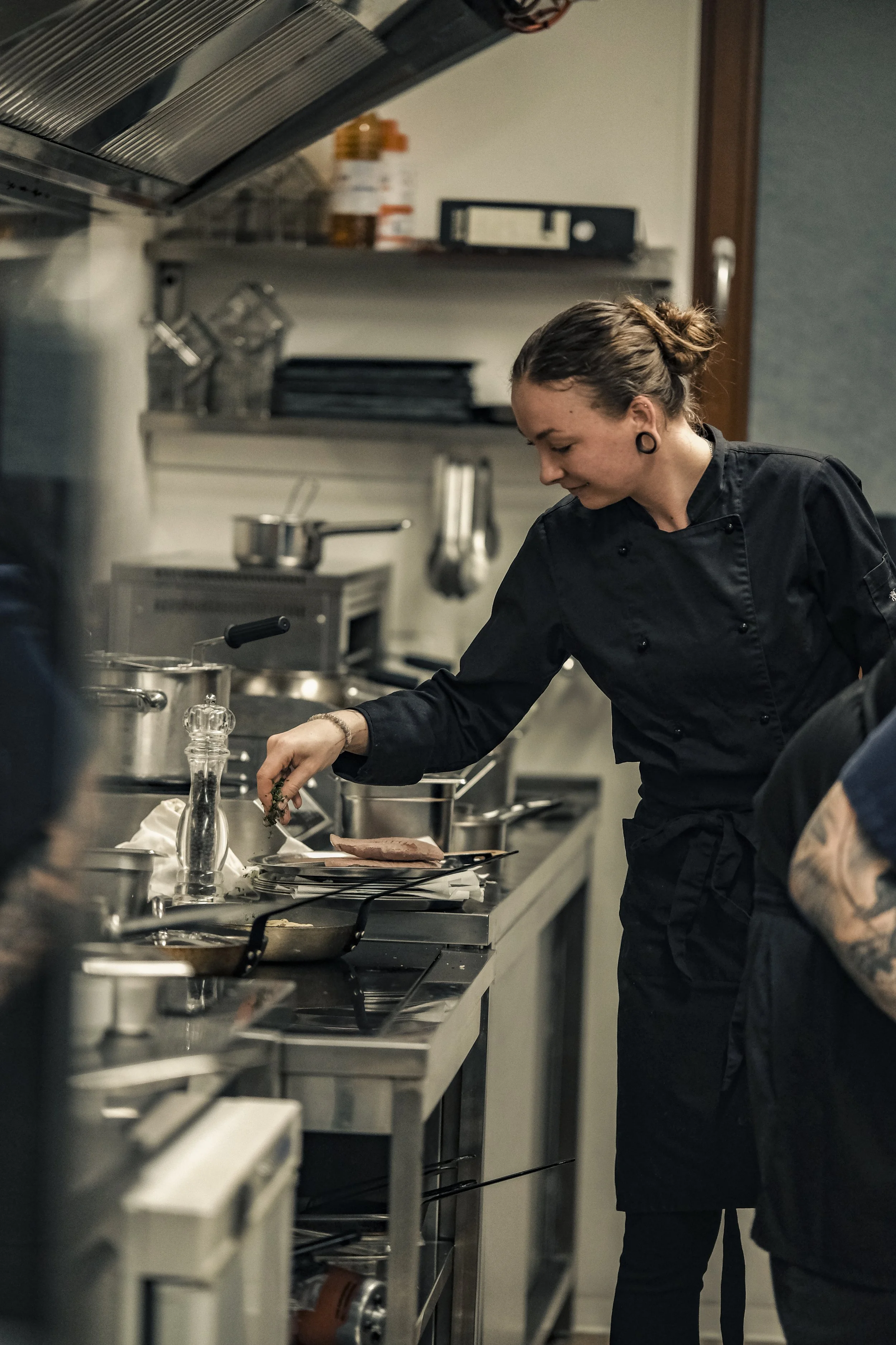 A female chef wearing a black chef's coat is preparing food in a commercial kitchen, seasoning a piece of meat on a plate.