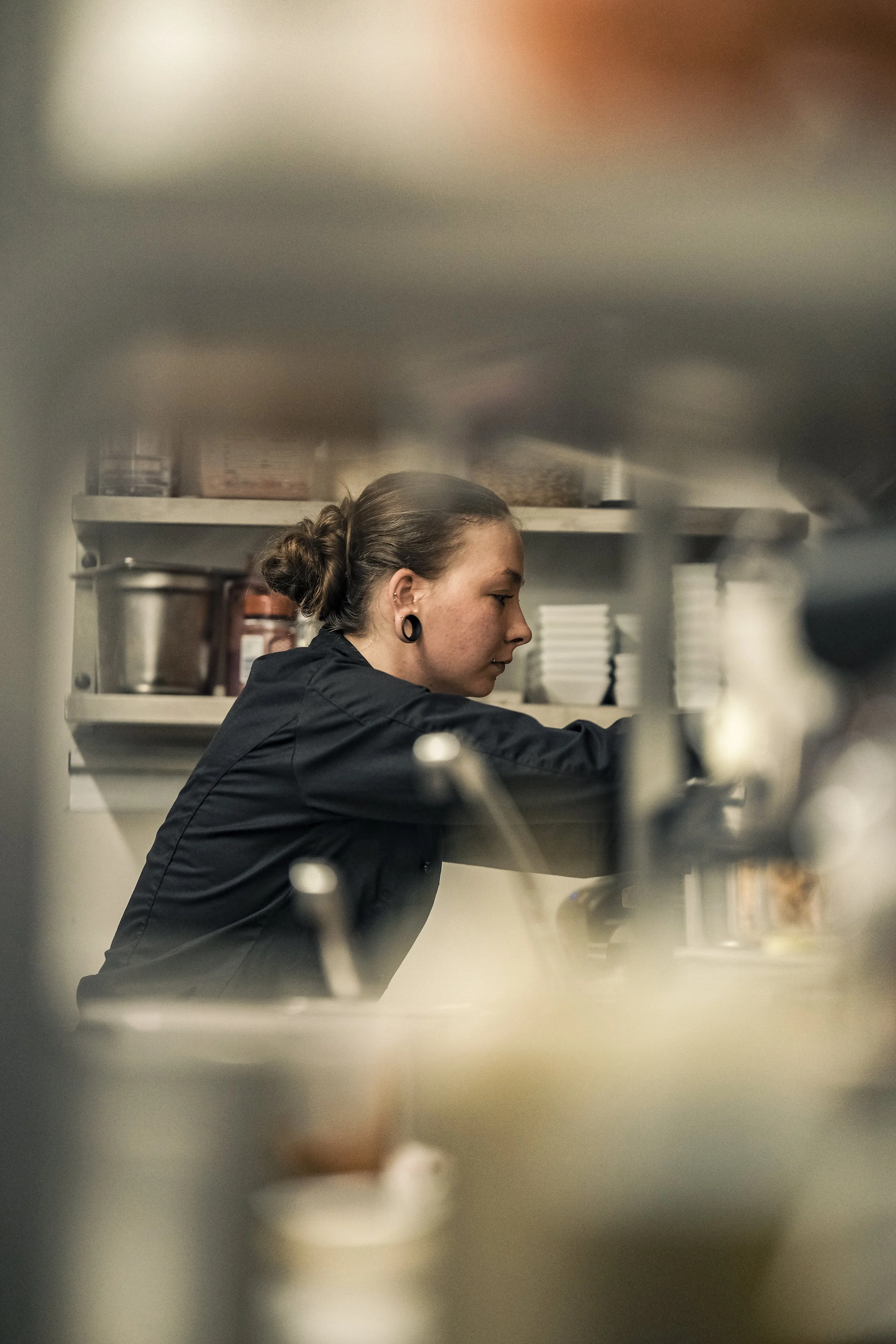 A woman with dark hair tied back, wearing black earrings and a black shirt, reaching into a kitchen shelf surrounded by various containers and ingredients.