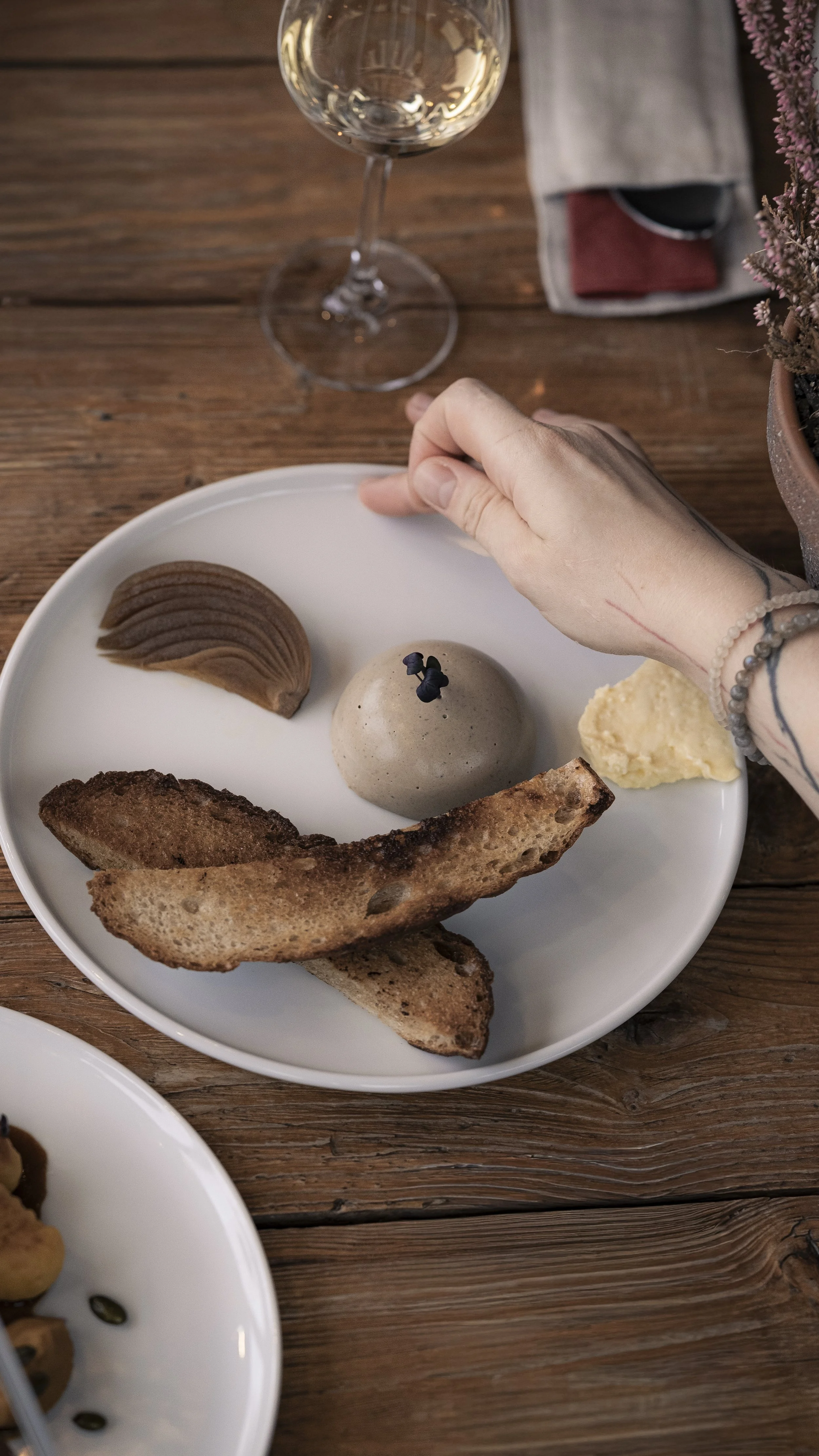 A white plate with bread slices, a scoop of sorbet topped with black berries, a prawn-shaped cookie, and dollops of cream on a wooden table with a glass of white wine in the background.