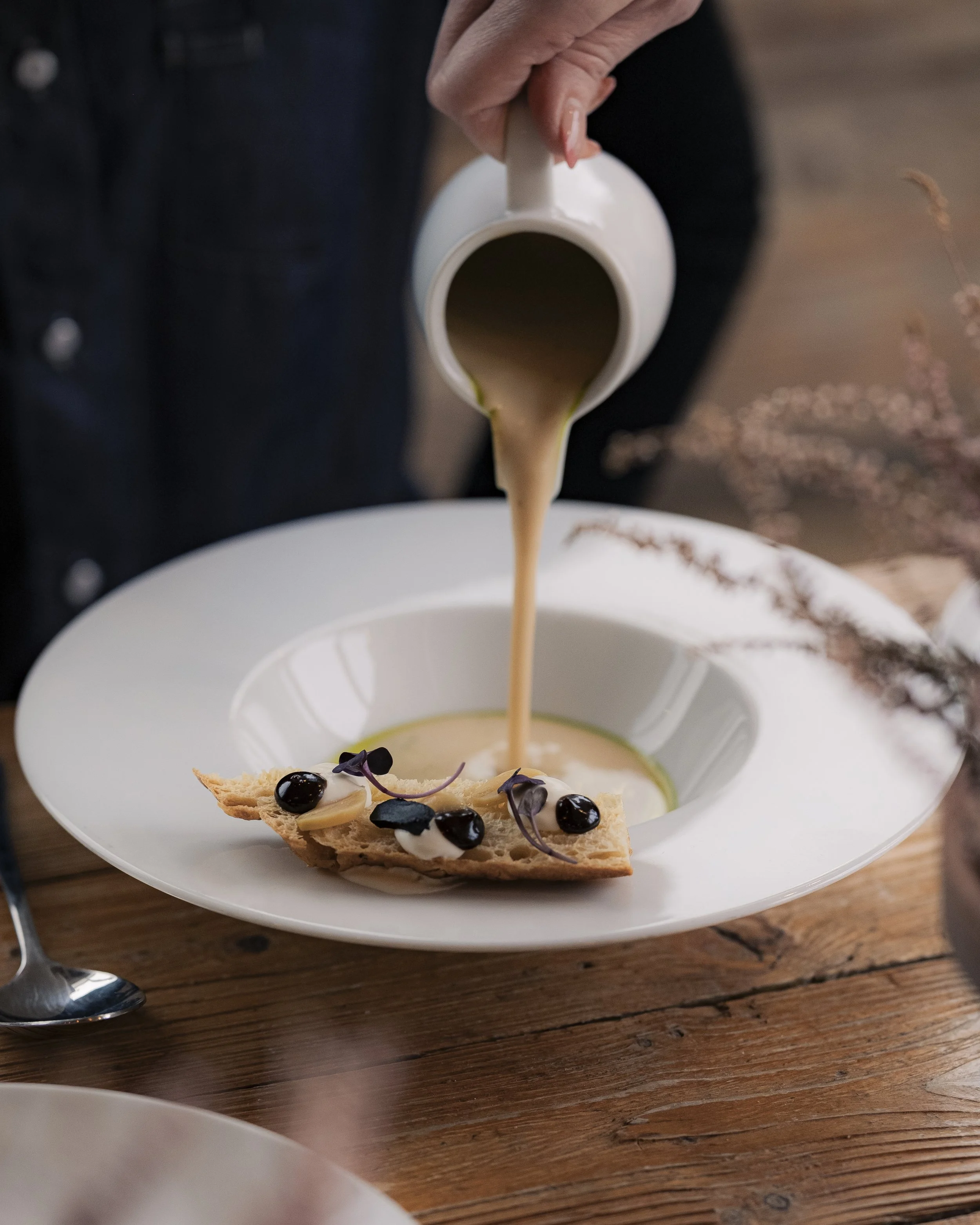 Pouring creamy soup from a white pitcher into a bowl with a bread piece and black grapes, on a wooden table.