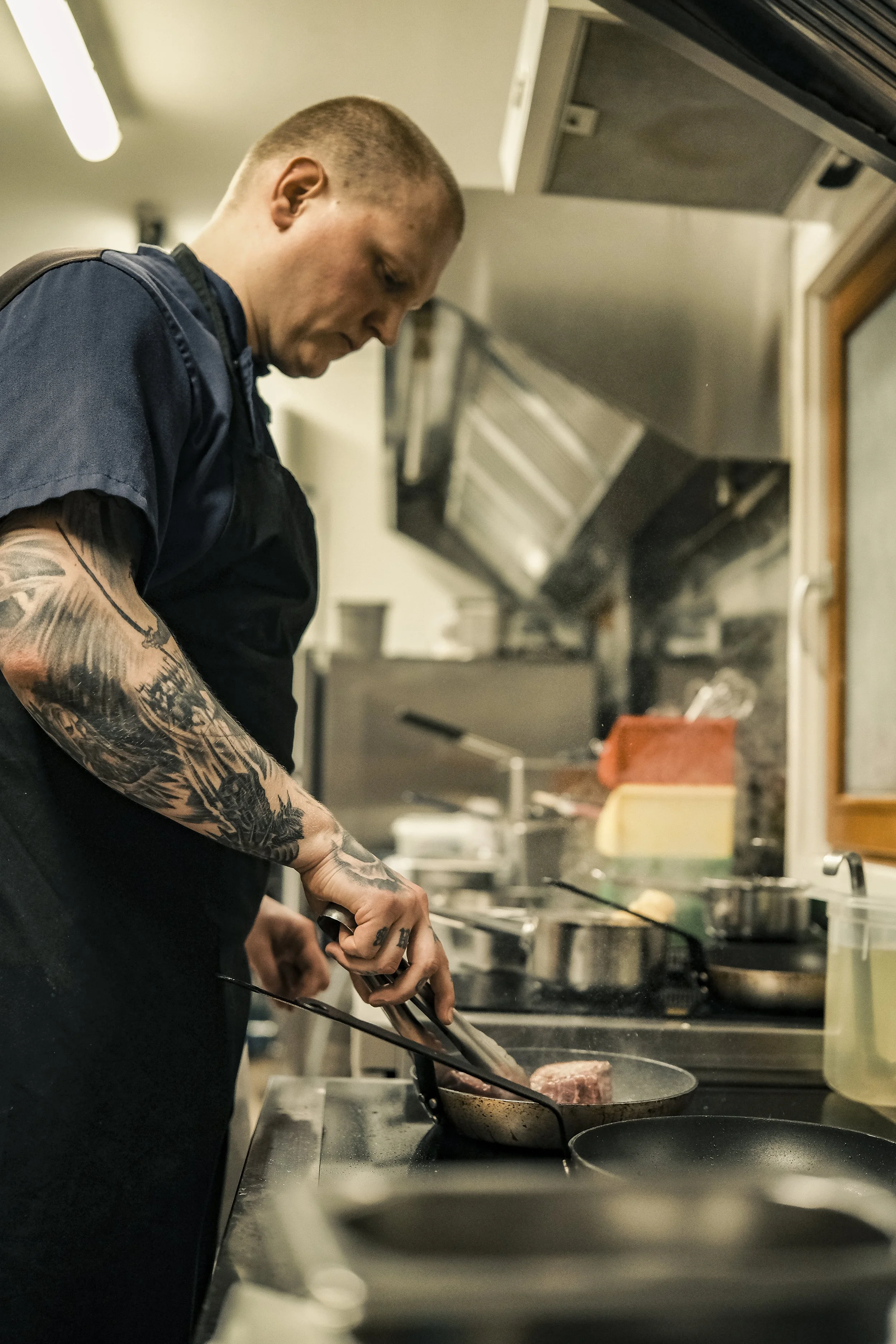 A chef with tattoos on his left arm is cooking steak in a pan on a stove in a professional kitchen.