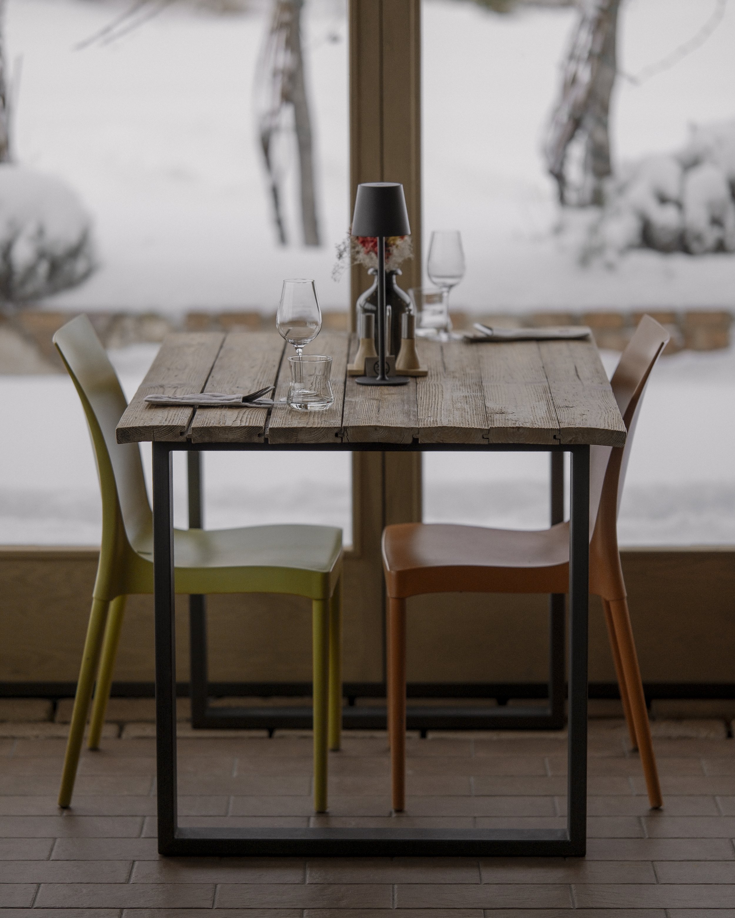 A cozy dining table set for two near a snowy window, with glassware, a black lamp, and minimalist decor.