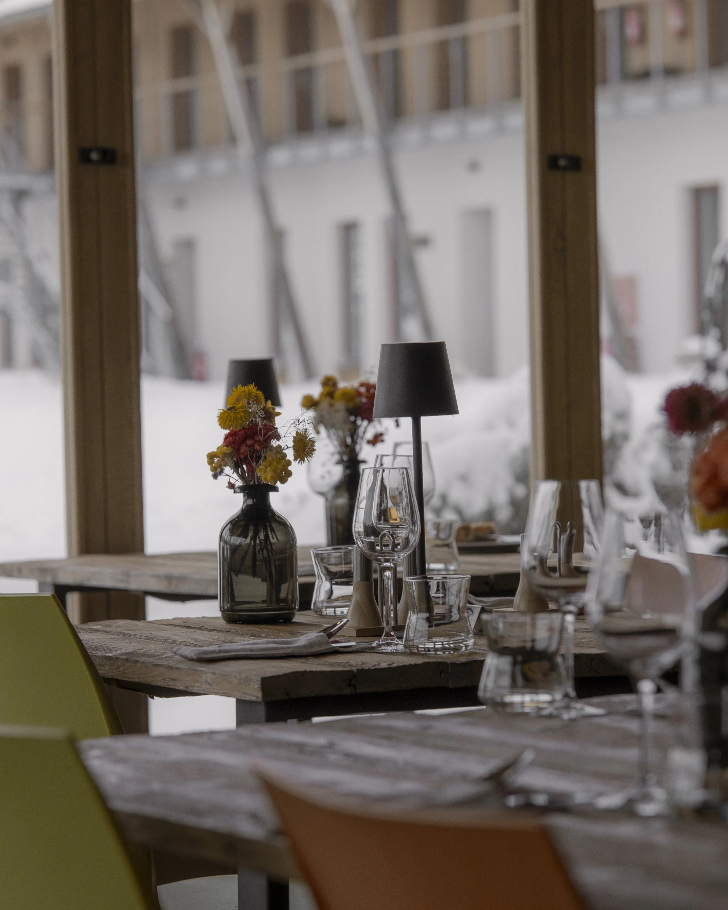 A cozy dining area with wooden tables, colorful chairs, flower vases, wine glasses, and lamps, overlooking a snowy outdoor scene through large windows.