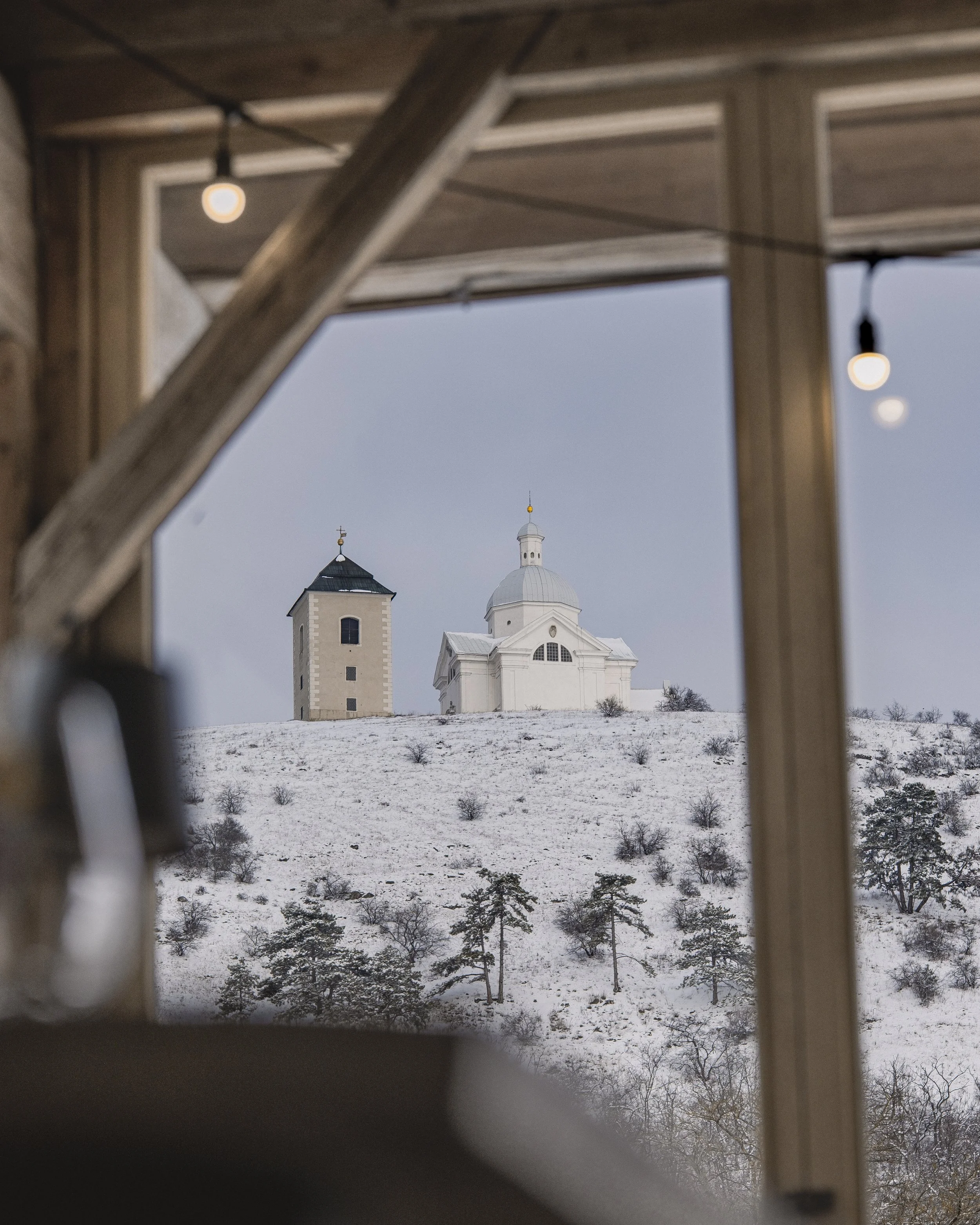 View of a snow-covered hillside with a white church and a small tower seen through a wooden-framed window or opening.