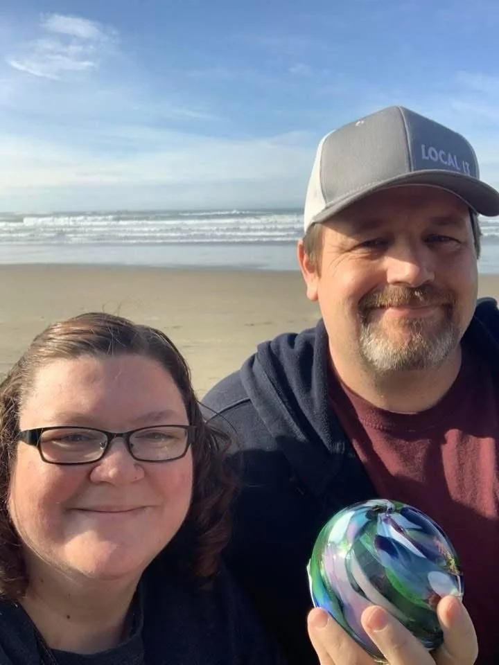 A smiling woman with glasses and a man with a beard and cap taking a selfie on a beach with sand and ocean waves in the background.