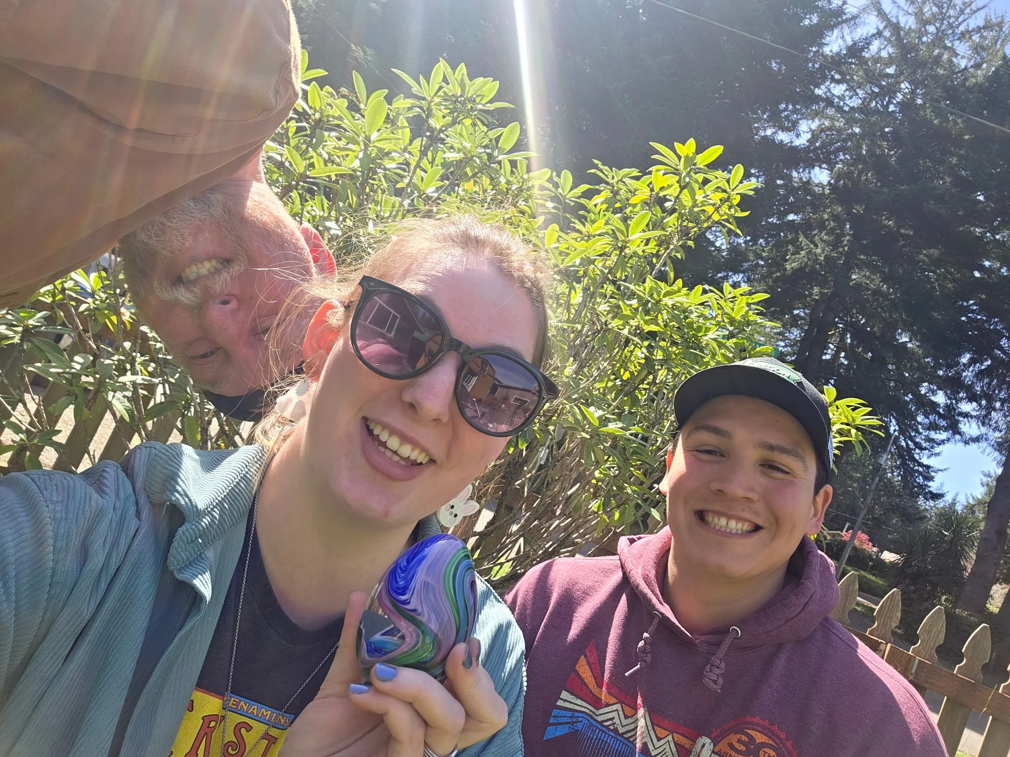 Four people are taking a selfie outdoors in sunlight, with trees and a wooden fence behind them. Two women and two men are smiling at the camera. One woman is holding a colorful glass art piece.