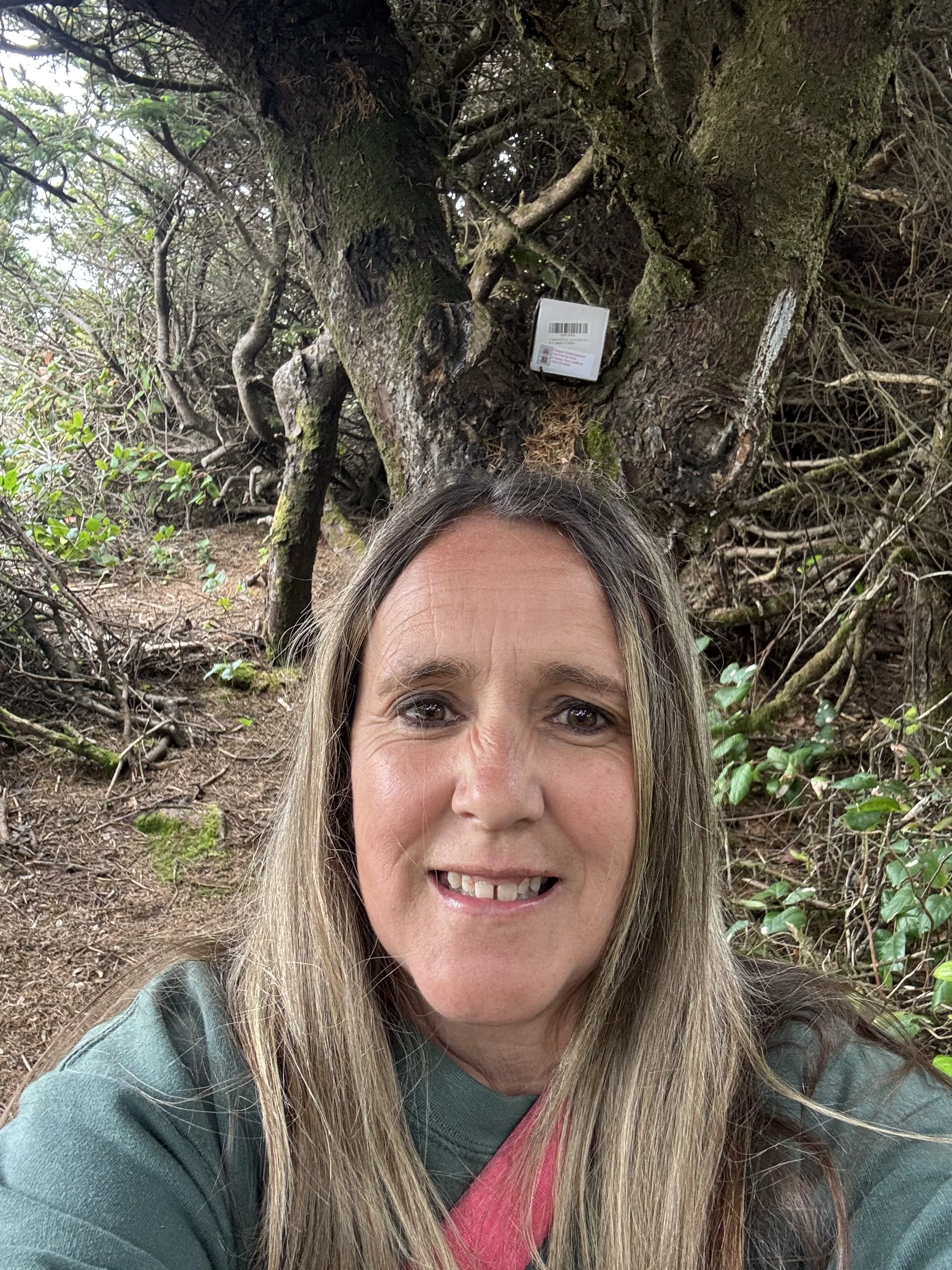 A smiling woman with long blonde hair taking a selfie outdoors in front of a large, moss-covered tree with twisted branches and moss. The woman is wearing a green jacket and has a camera strap around her neck.