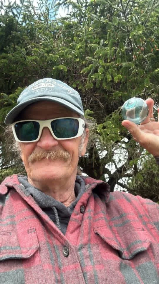 A man with a mustache wearing sunglasses, a baseball cap, and a plaid shirt holds a glass paperweight outdoors in front of green bushes.