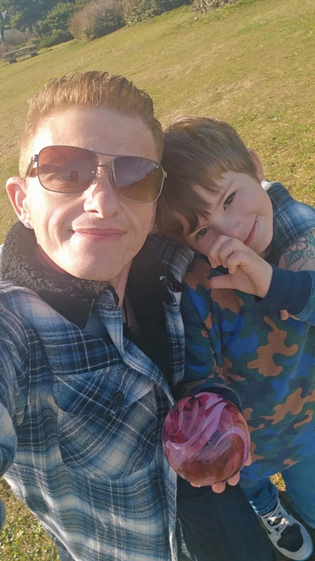A woman and a young boy taking a selfie outdoors on a sunny day. The woman is wearing sunglasses and a plaid jacket, smiling. The boy, wearing a camouflage hoodie, is smiling and making a heart shape with his hand. The background is a grassy field with trees.