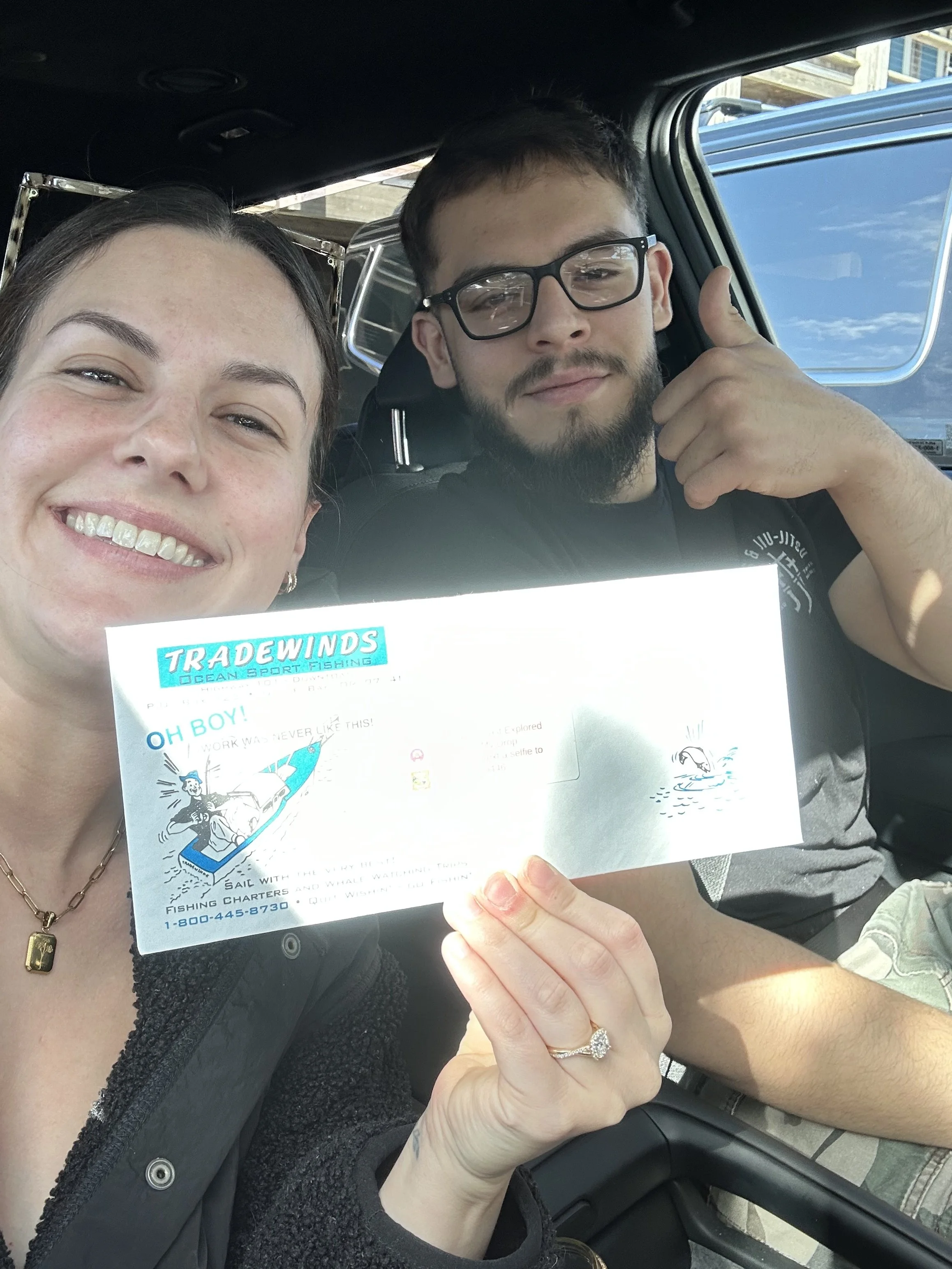Two people, a woman and a man, taking a selfie inside a vehicle. The woman is smiling and holding a flyer for Tradewinds Ocean Sport Fishing, which features a cartoon of a person fishing from a boat. The man is wearing glasses and making a thumbs-up gesture. The background shows part of the vehicle's interior and an outdoor scene through the window.