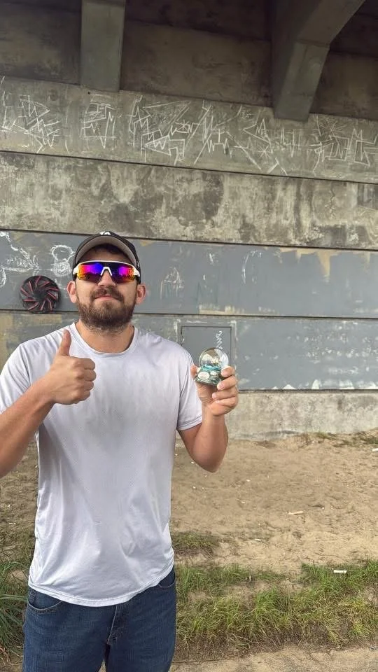 Man wearing sunglasses, a white t-shirt, and a cap, holding a round object in his right hand and giving a thumbs-up with his left hand under a bridge with graffiti and concrete panels.