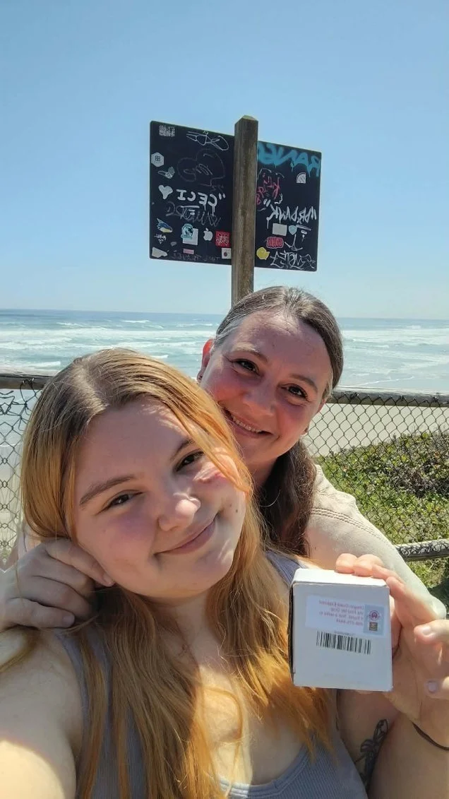 Two women smiling at the beach, one holding a parking pass or ticket, with a fence and ocean behind them, and graffiti-covered signs above.