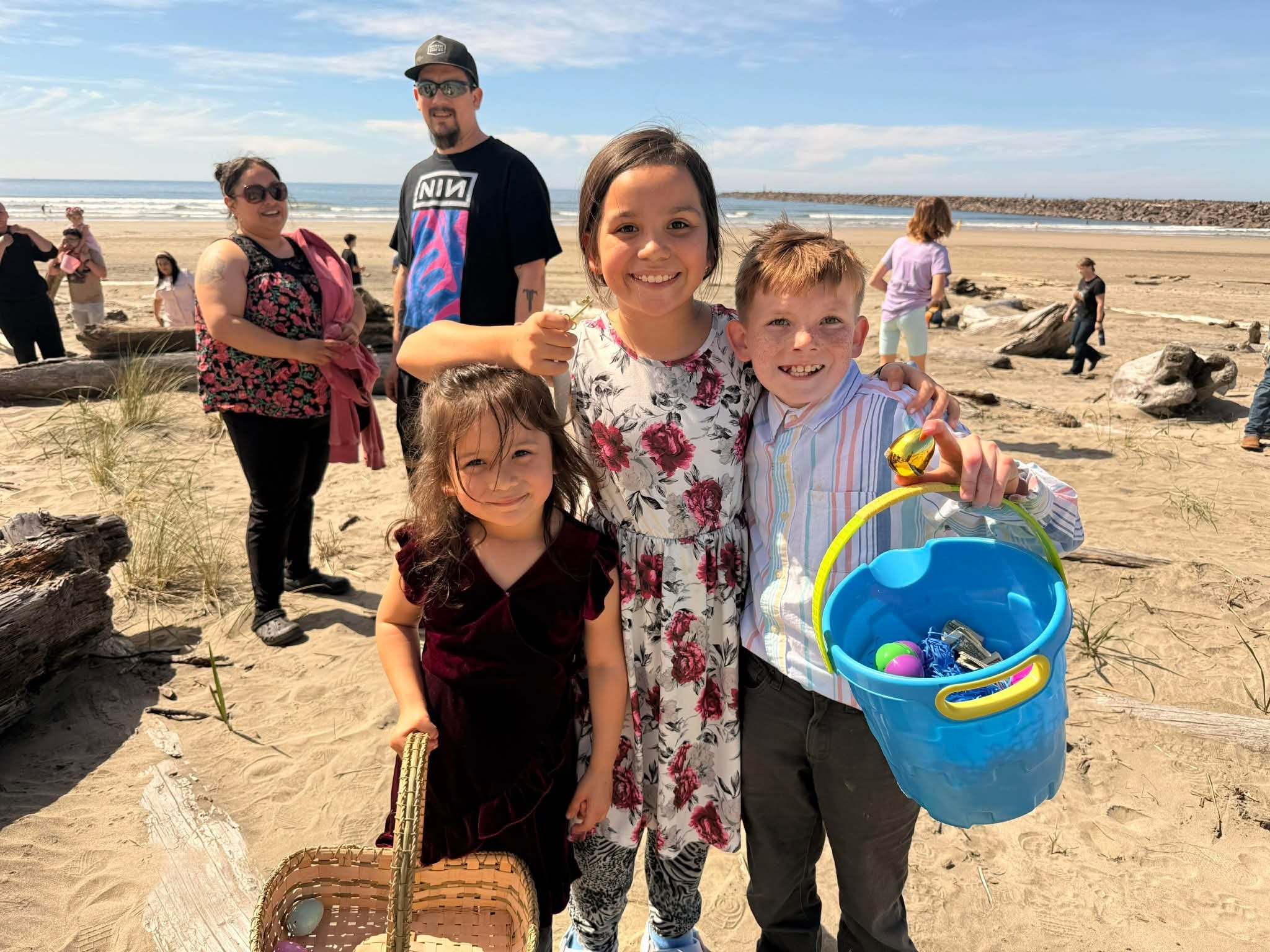 People gathered on a beach, smiling and holding buckets with colorful Easter eggs, with the ocean and a rocky jetty in the background.