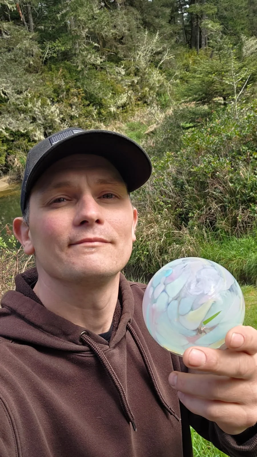 A man wearing a brown hoodie and a black cap is holding a colorful glass marble in an outdoor setting with green trees and shrubs in the background.
