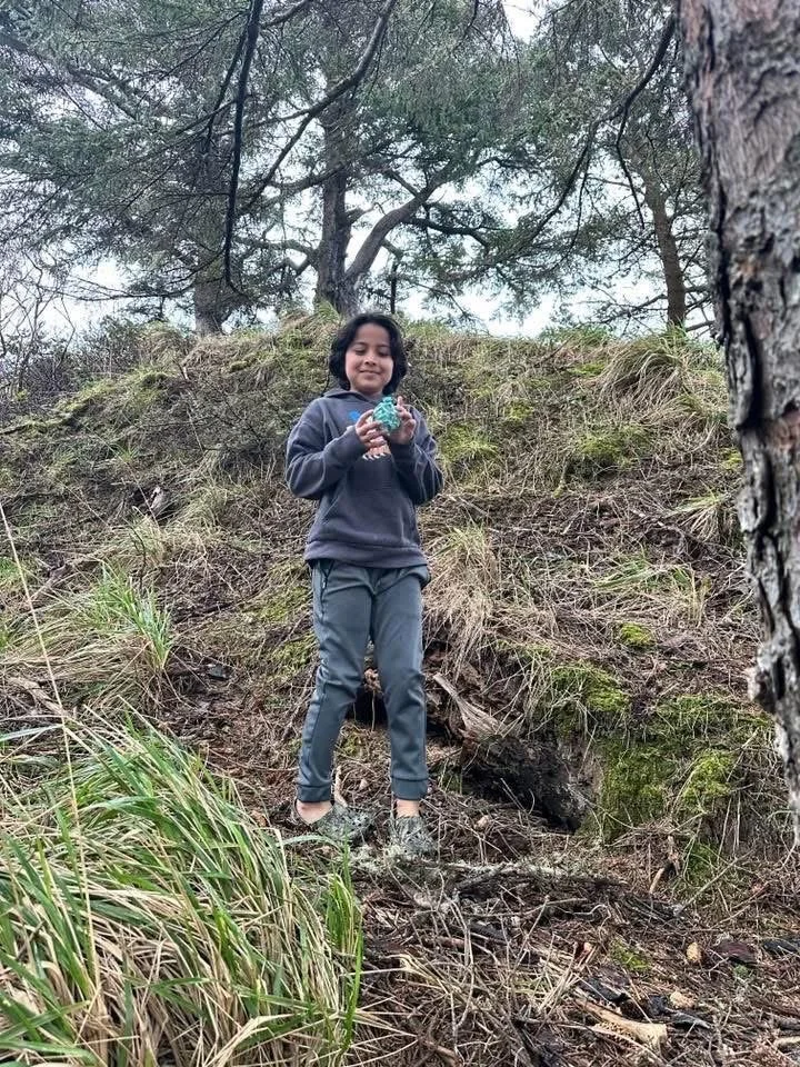A girl standing on a hill holding a small toy, surrounded by trees and grass.