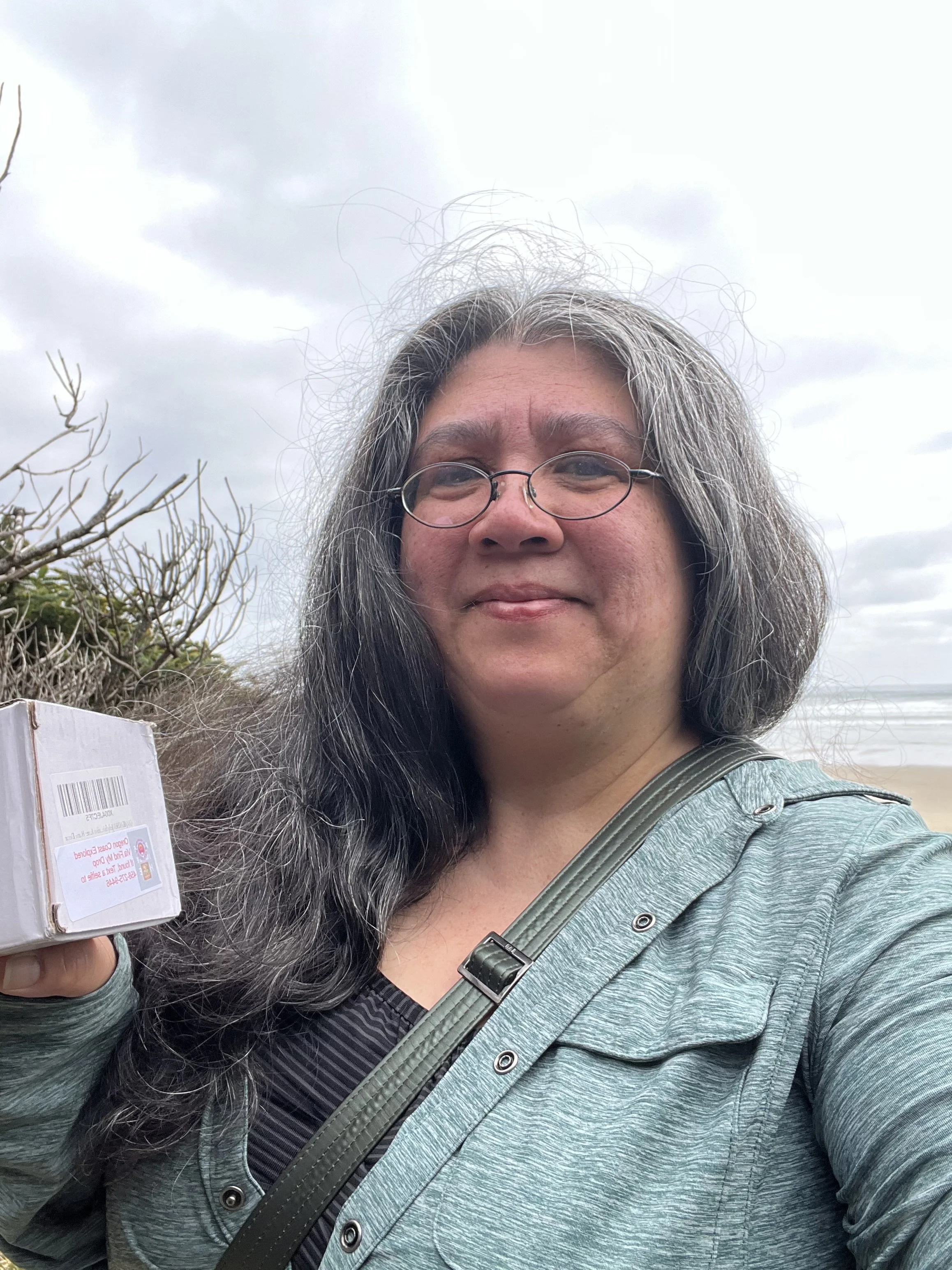 A woman with long gray hair, glasses, and a green jacket standing outdoors near the beach, holding a box of cereal, with cloudy sky and beach in the background.