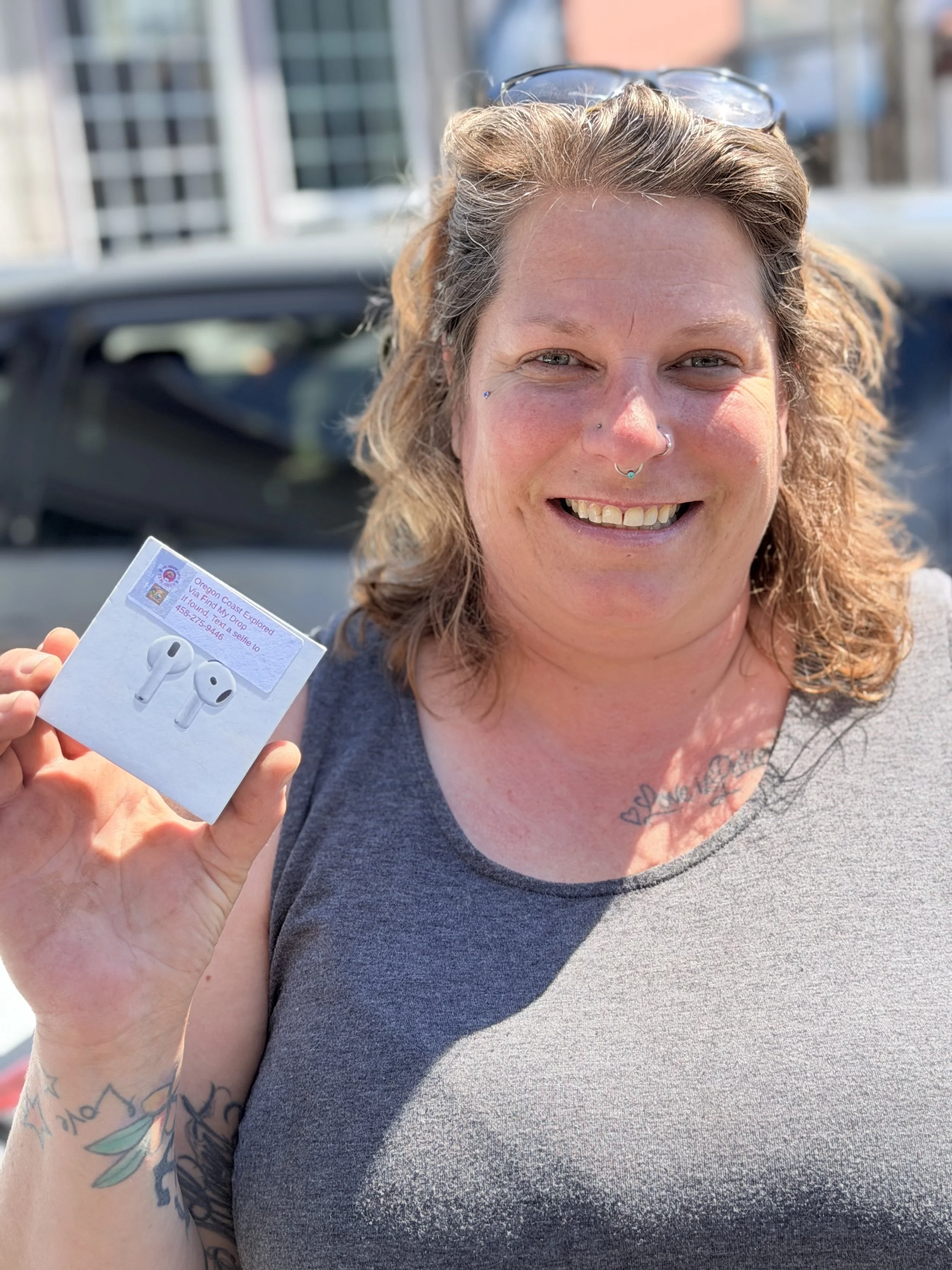 A woman with curly hair, tattoos on her arm, and multiple facial piercings, smiling outdoors, holding a pair of wireless earbuds and their box.