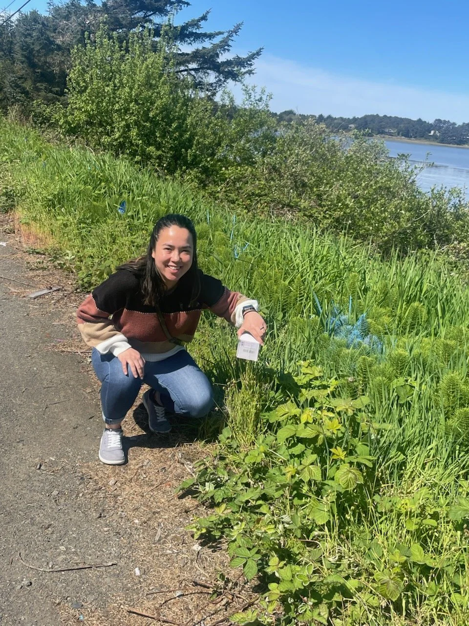 A woman crouching by a lush green roadside with a river in the background on a sunny day.