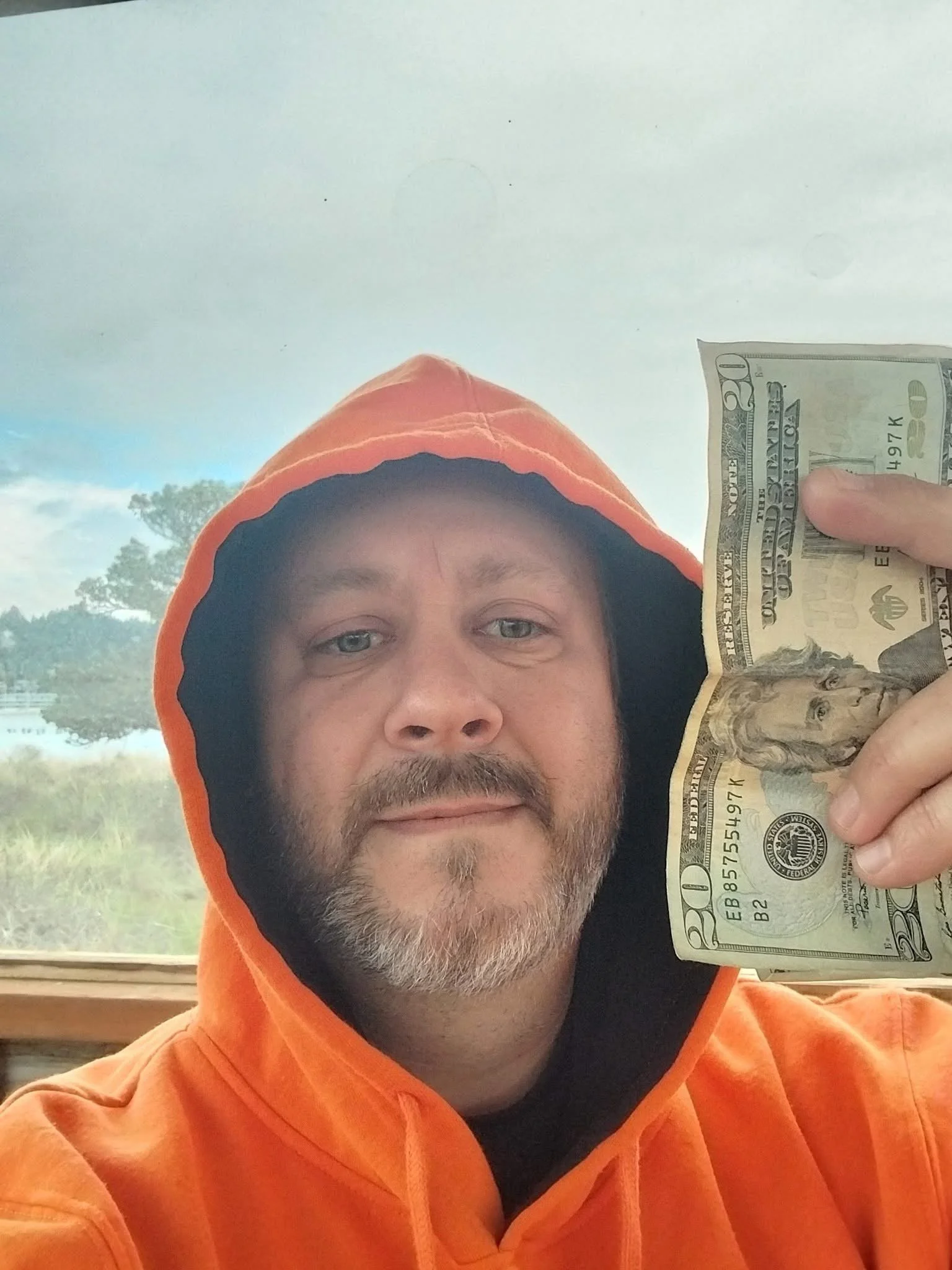A man with a beard wearing an orange hoodie and black shirt, holding a twenty-dollar bill near his face, in front of a window with a view of trees and a cloudy sky.