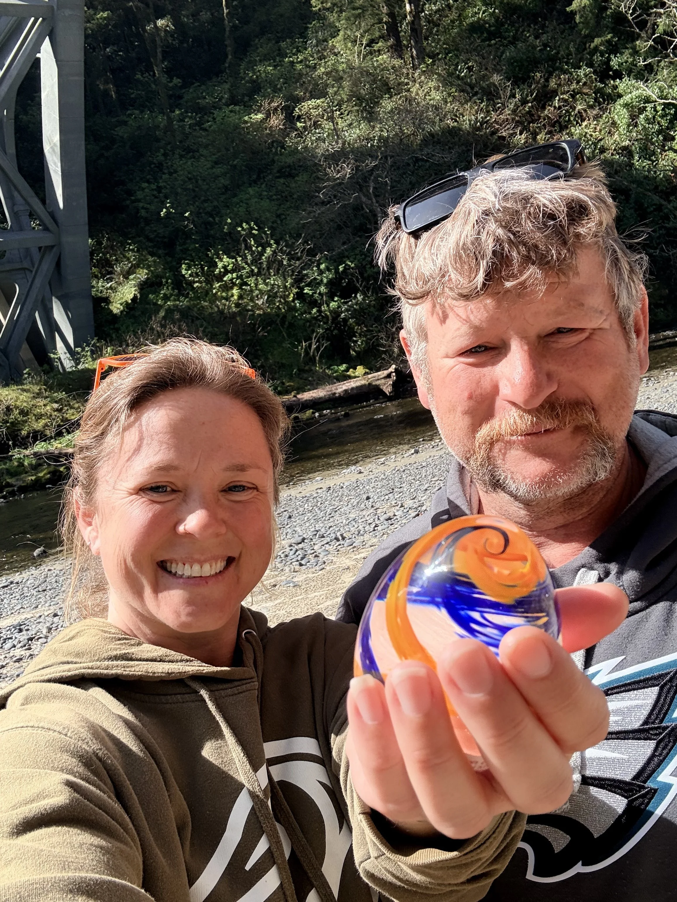 A smiling woman and a man smiling outdoors by a river, with a bridge in the background. The woman has blonde hair, the man has curly gray hair and a beard, and they are holding a colorful glass object towards the camera.