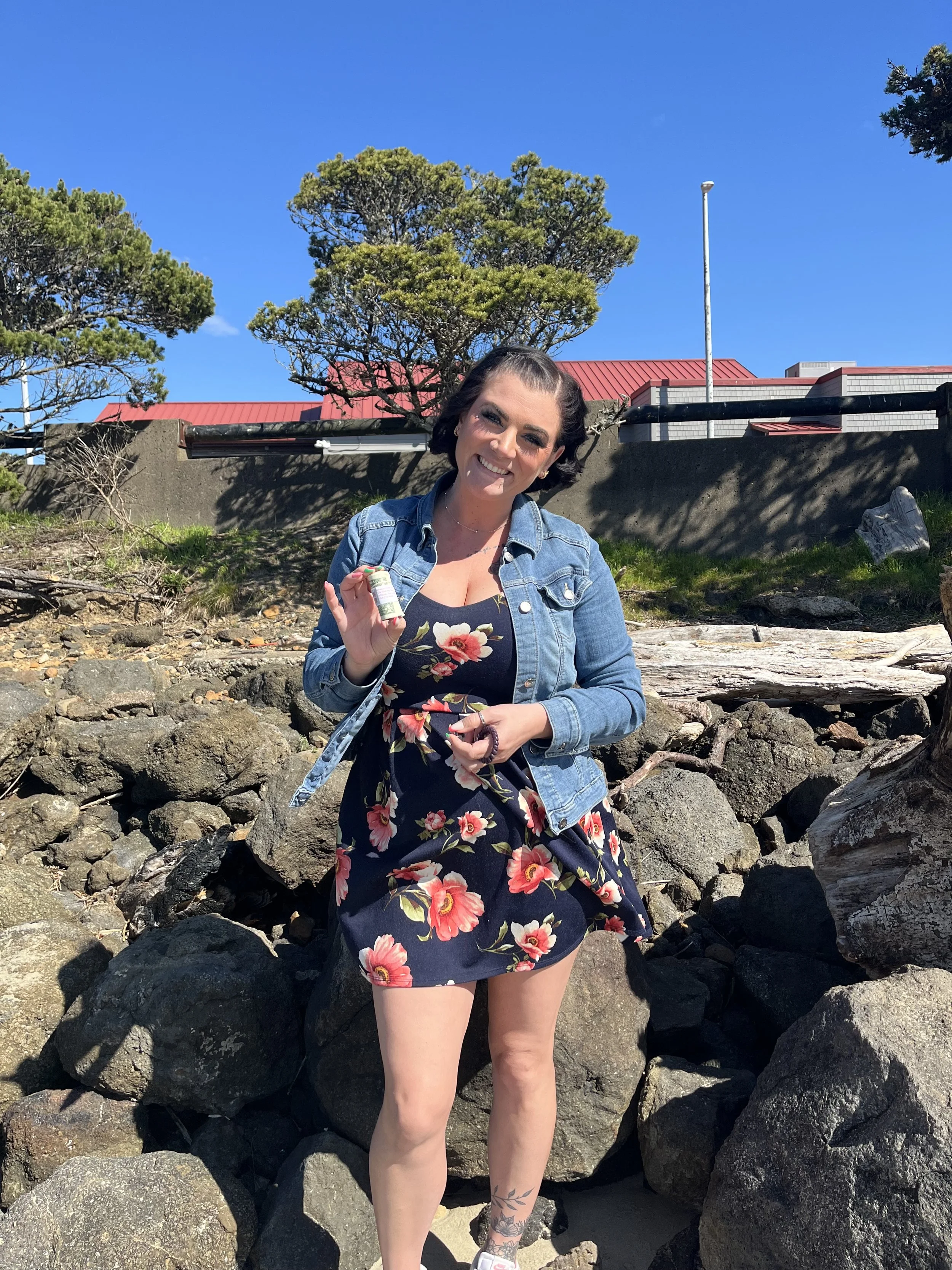 A woman with short dark hair and tattoos on her legs, wearing a floral dress and denim jacket, standing on rocks near the ocean, holding a small bottle, smiling at the camera under clear blue sky.