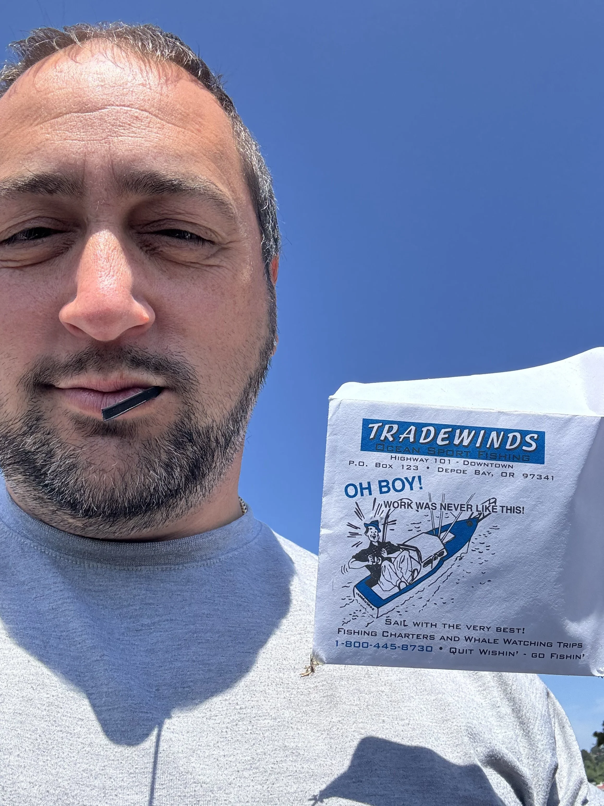 A man taking a selfie outdoors with a bright blue sky in the background, wearing a light-colored shirt and smiling with his eyes partially closed. Beside him is a flyer for Tradewinds Ocean Sport Fishing, featuring a cartoon image of a man fishing from a boat, holding a fish, with text advertising fishing trips.