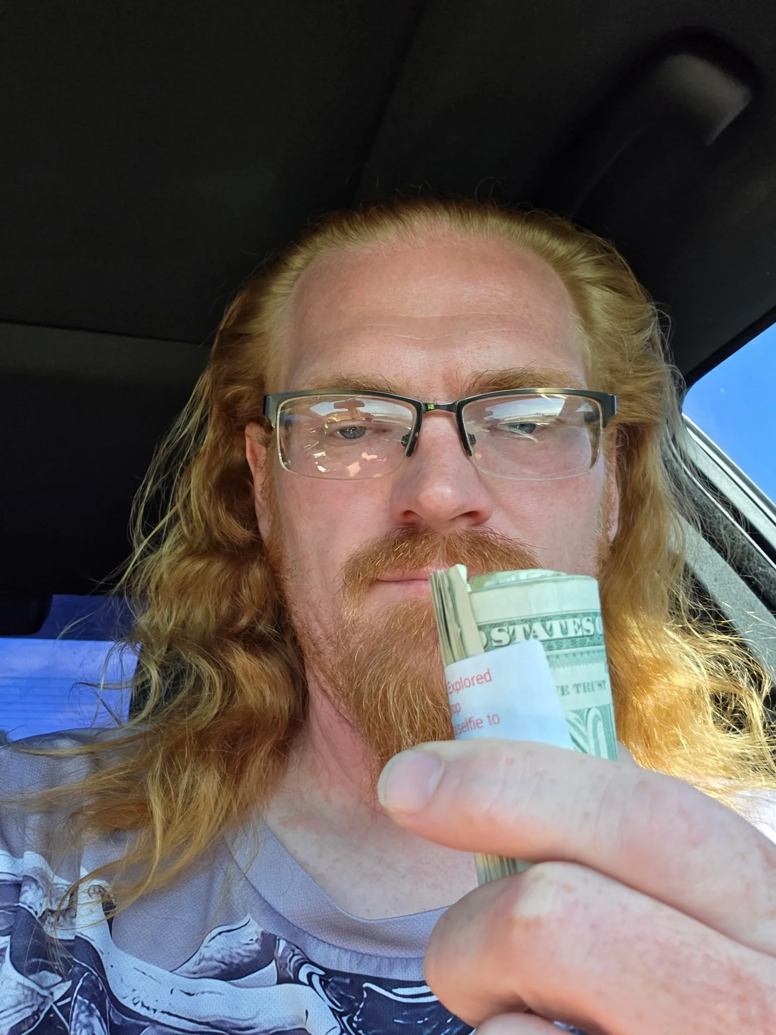 A man with long red hair and glasses holding a small bundle of cash in his hand inside a vehicle.