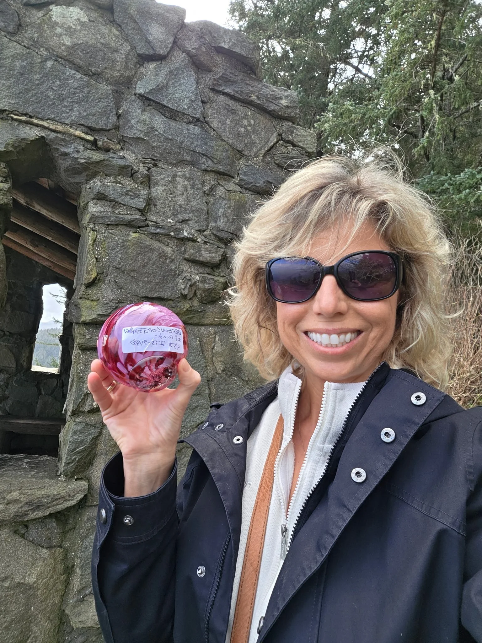 A woman with blonde hair wearing black sunglasses and a black jacket taking a selfie outdoors, holding a pink glass ornament with a note attached, standing in front of a stone structure with greenery in the background.