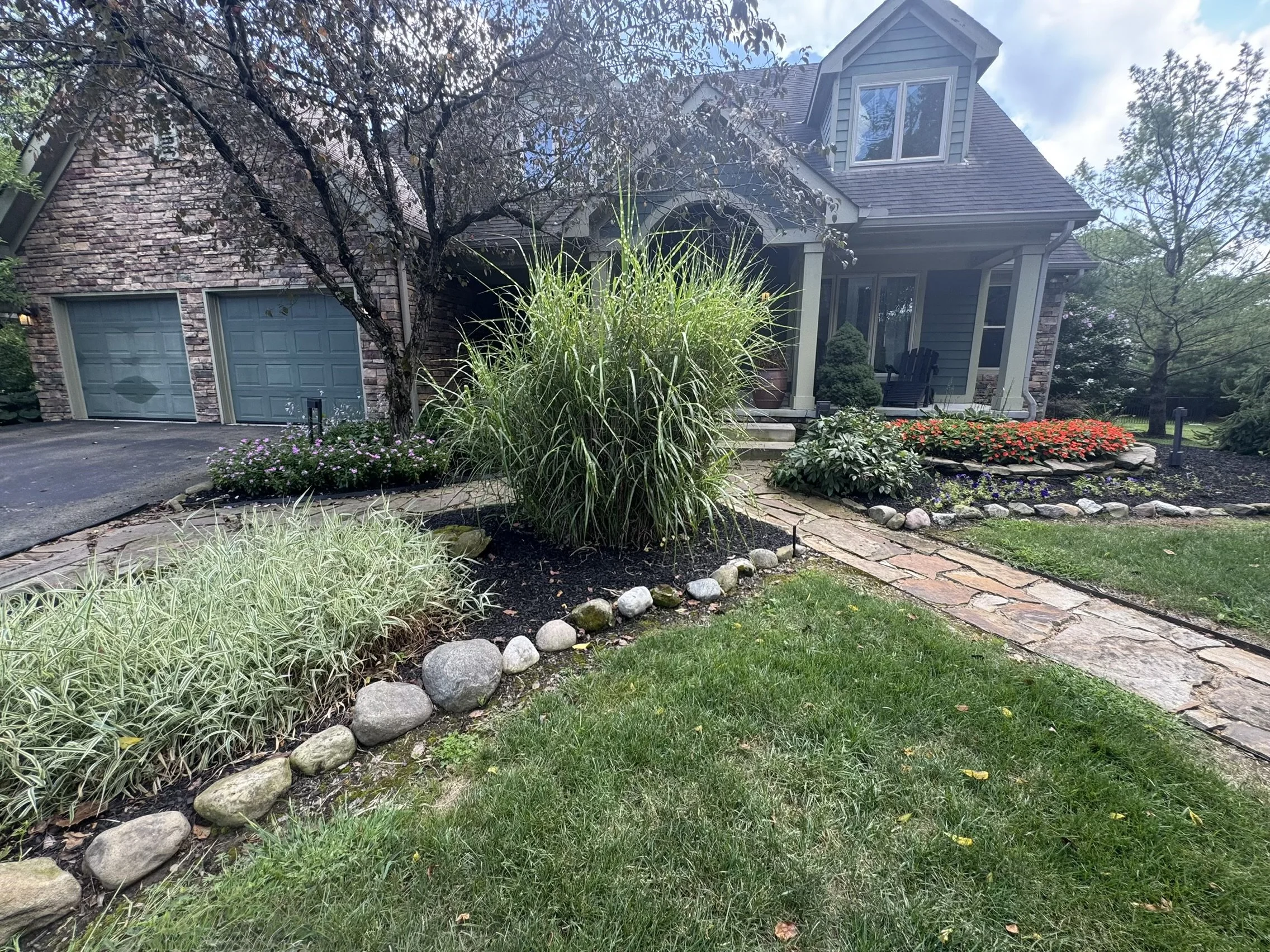 Front yard of a house with a stone walkway, lush greenery, colorful flowers, and a porch with Adirondack chairs.
