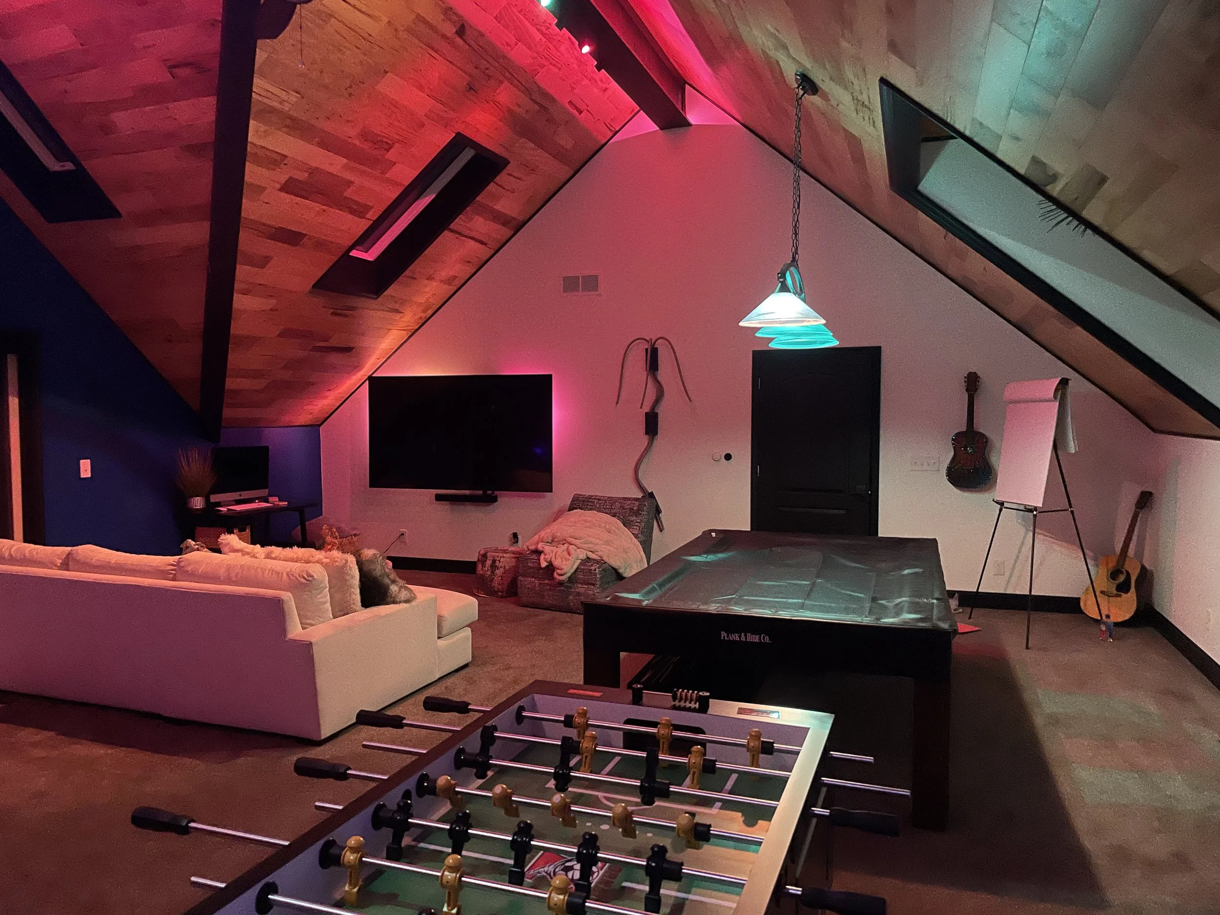 Living room with sloped wooden ceiling, TV, white sofa with pillows, foosball table, guitars, flip chart, and colorful lighting.