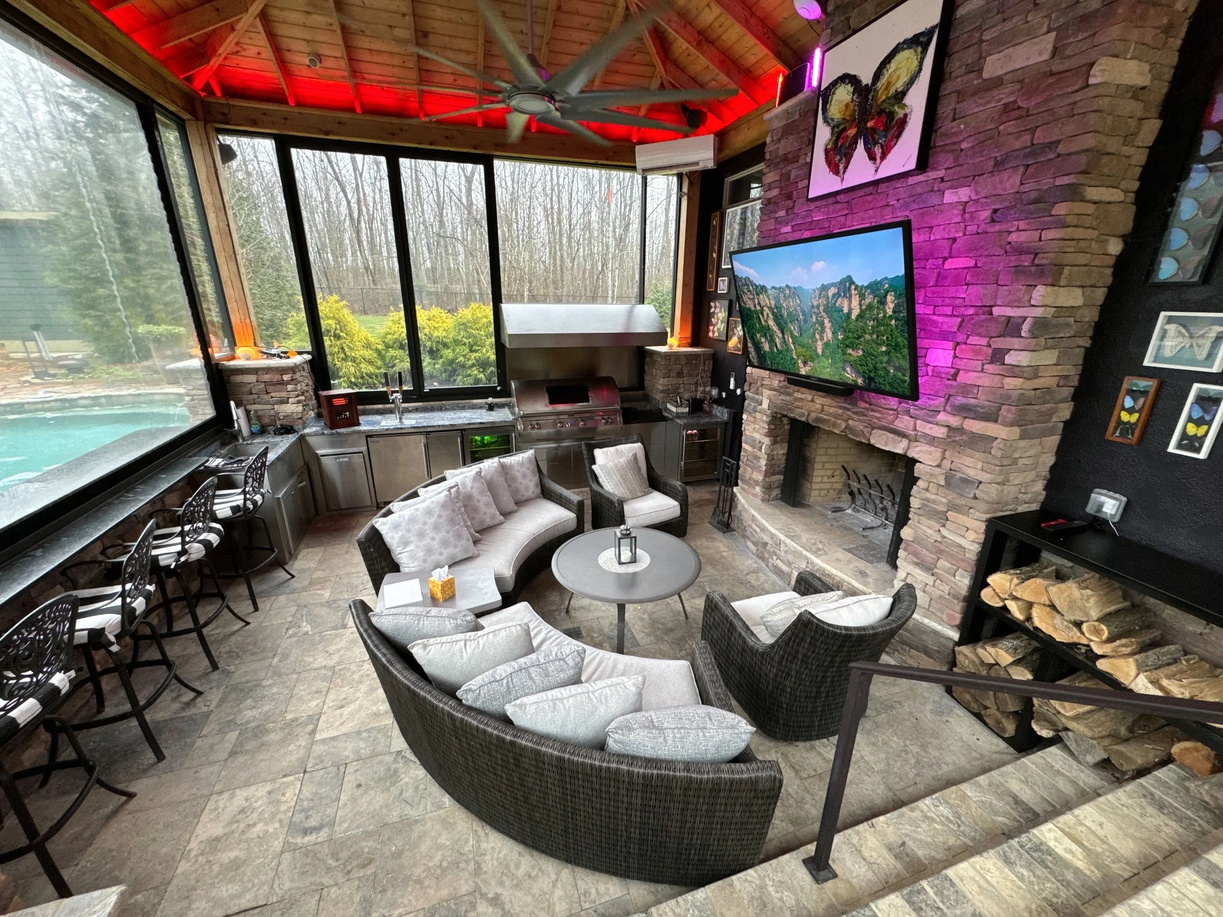 Indoor living space with a large window, a stone fireplace with a mounted TV, a curved sofa with pillows, a round coffee table, chairs, a bar with chairs, and a view of trees outside.