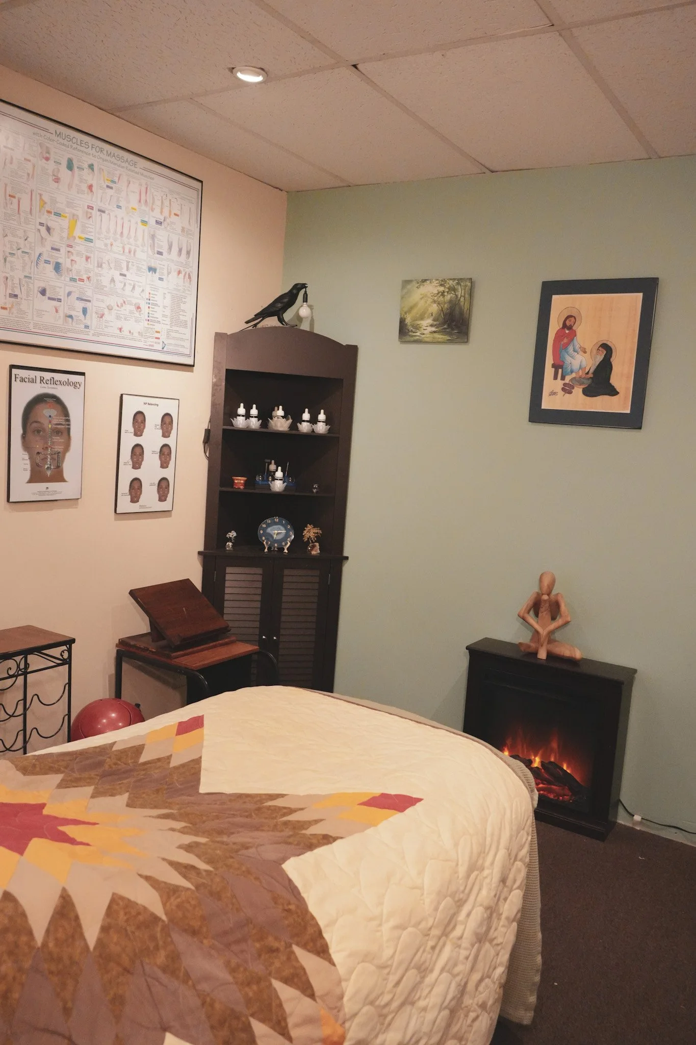 A cozy room with a bed covered in a quilt with a star design, a small electric fireplace, framed artwork on the walls, a black shelving unit with various small objects, and a stool with a book on it.