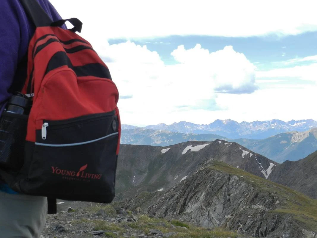 A person hiking in a mountainous landscape with a red and black Young Living backpack, mountain peaks in the background, and partly cloudy skies.