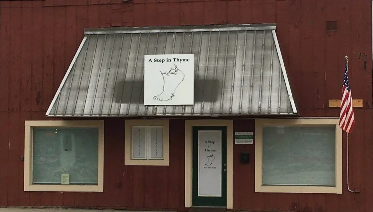 Front view of a red building with a metal roof, featuring a sign that reads "A Step in Thyme" and includes a drawing of a foot with a thyme plant. The building has two large windows with beige frames, a door with the same sign, and an American flag on a pole to the right.