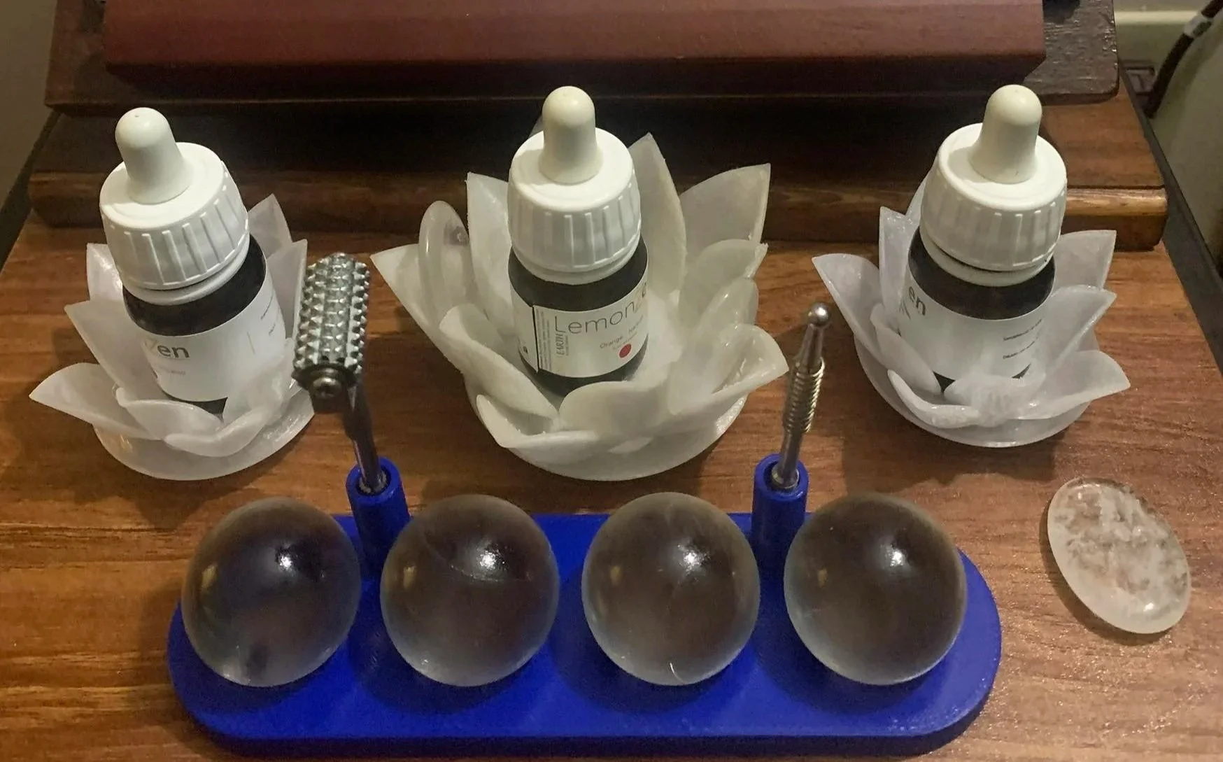 Three bottles of essential oils in flower-shaped white bowls, two small metal tools, four black spherical stones, and a white stone on a wooden surface.