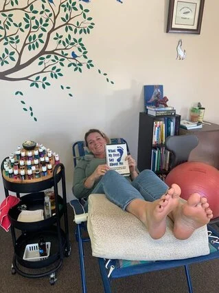 Woman lying on a bed with her feet up, holding a book, in a room with a wall mural of a tree, a bookshelf, a therapy ball, and a small table with jars and other items.
