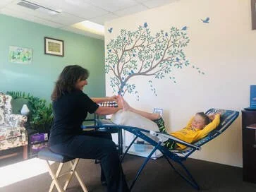 A woman sitting at a table and reading to a young girl lying on a hammock in a colorful room with a wall mural of a tree and birds.