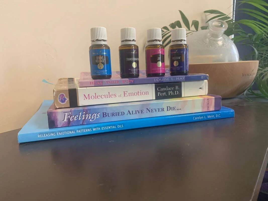 Four small bottles of essential oils are placed on top of three books. The titles of the books include "Molecules of Emotion," "Feelings Buried Alive Never Die," and "Releasing Emotional Patterns with Essential Oils." The background includes a plant and a glass container on a wooden base.