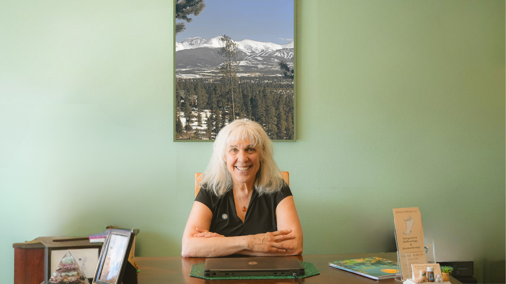 A smiling woman with white hair sitting at a wooden desk with a laptop in a room with light green walls; a landscape picture of snowy mountains and pine trees hangs on the wall behind her.