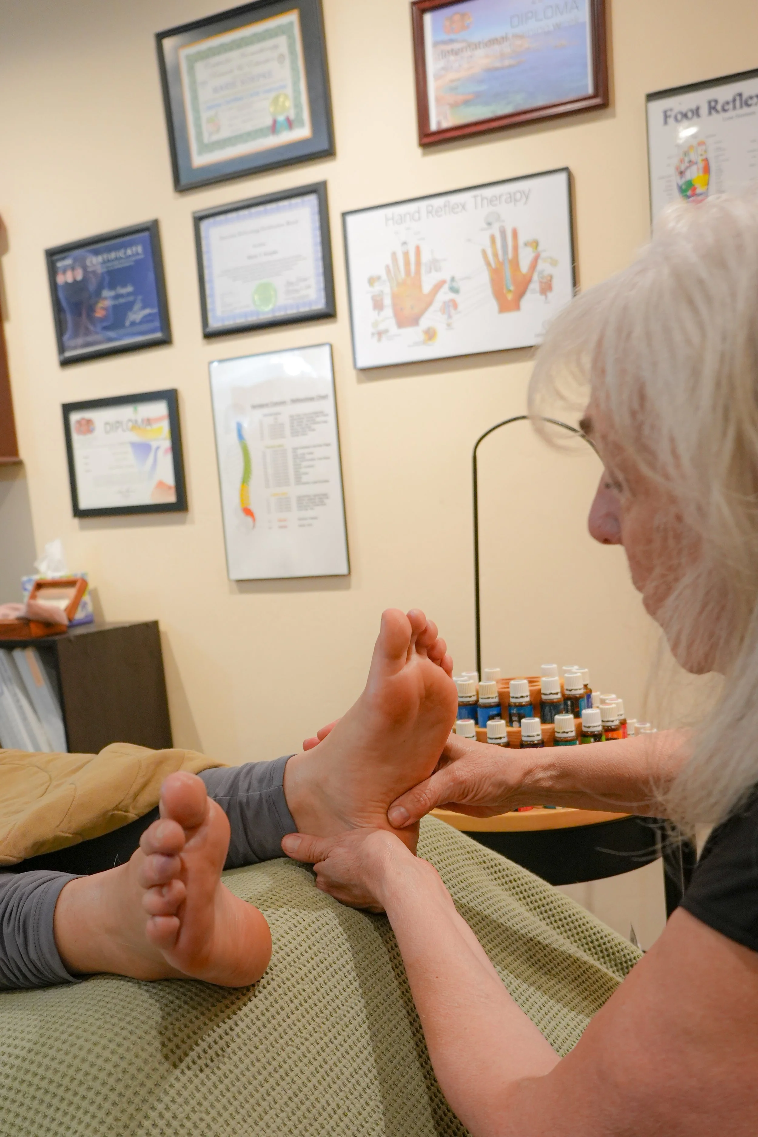 A person receives foot reflexology treatment from a therapist in a therapy room decorated with framed certificates and diagrams on the wall.