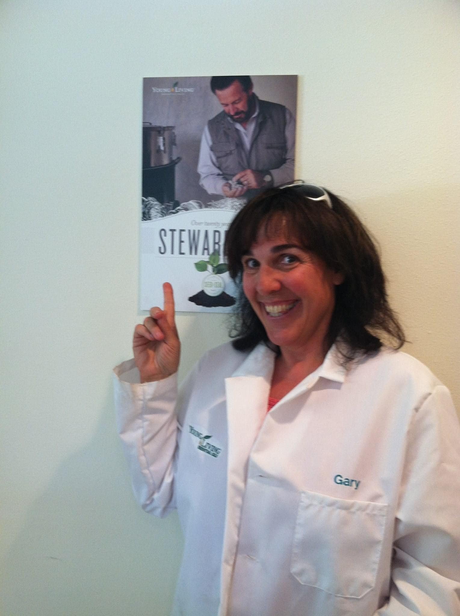 A woman wearing a white shirt with the name 'Gary' embroidered on it, smiling and pointing at a poster on the wall. The poster features a man in a vest and is labeled 'STEWAR.'