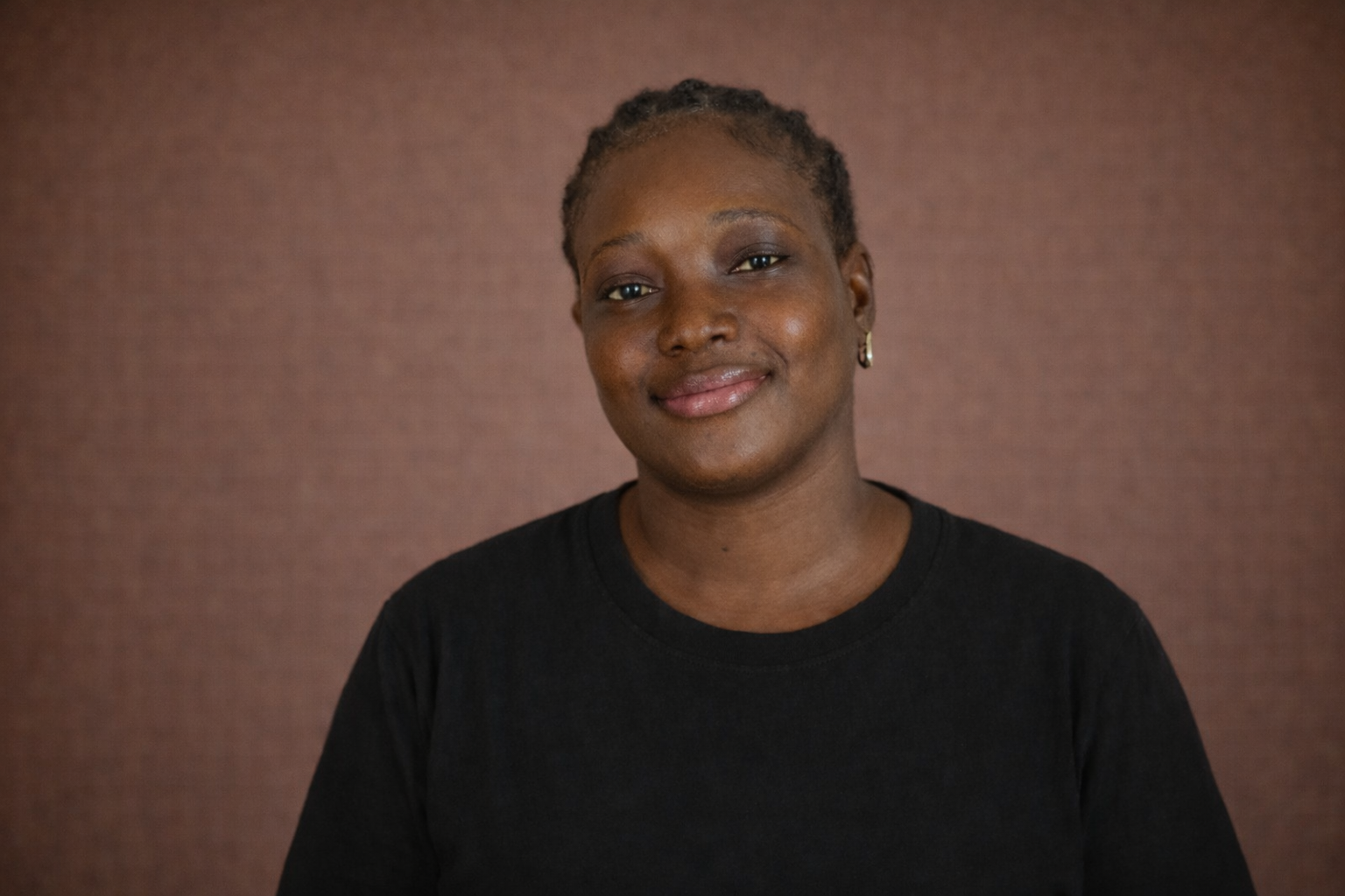 A woman with short hair smiling, wearing a black shirt and small hoop earrings, standing against a brown background.