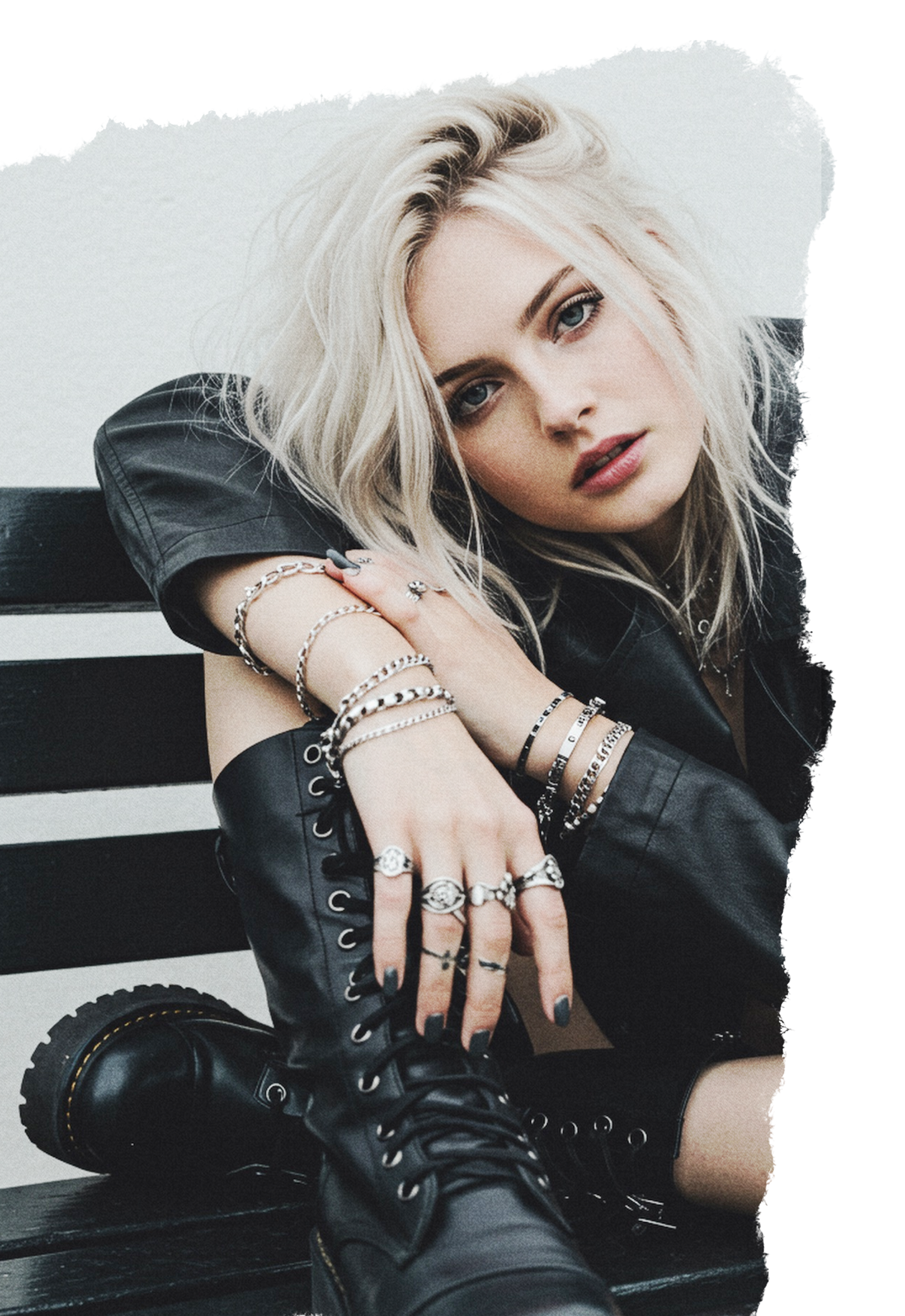 A young woman with blonde hair and blue eyes sitting on a black and white bench, wearing a black leather jacket, multiple silver rings and bracelets, and black lace-up boots, looking directly at the camera.