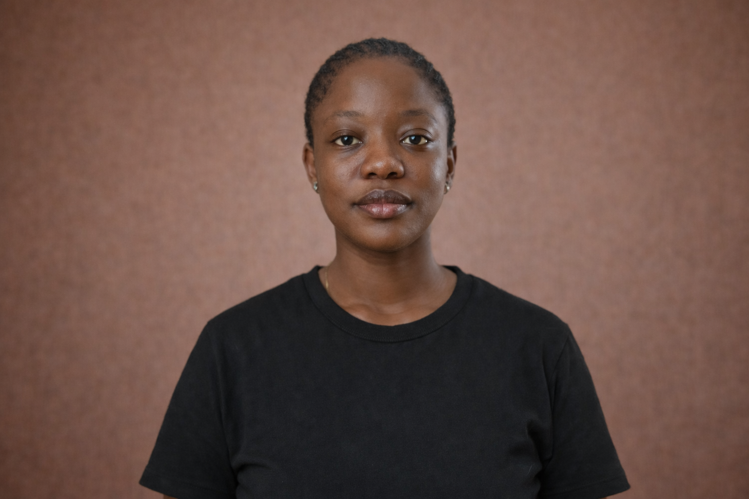 A young African American woman with braided hair, wearing a black t-shirt, standing against a plain brown background.