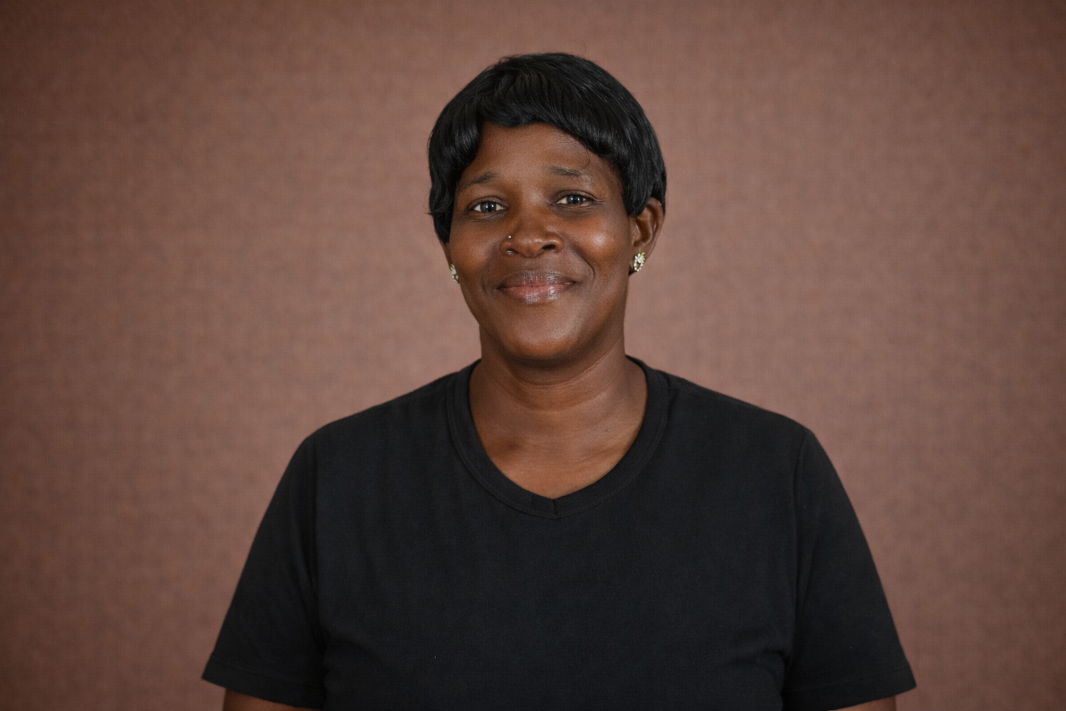 Headshot of a smiling woman with short black hair, wearing earrings and a black t-shirt, against a brown background.