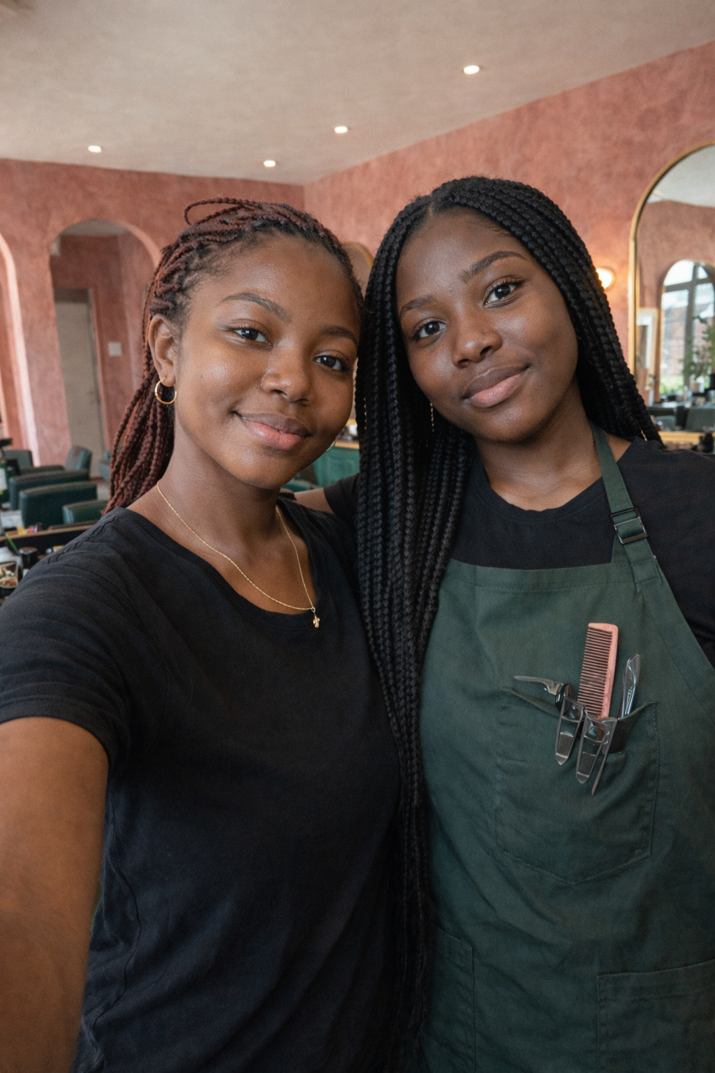 Two women taking a selfie in a restaurant. One has braided hair, and the other is wearing a green apron, holding hair-cutting tools.